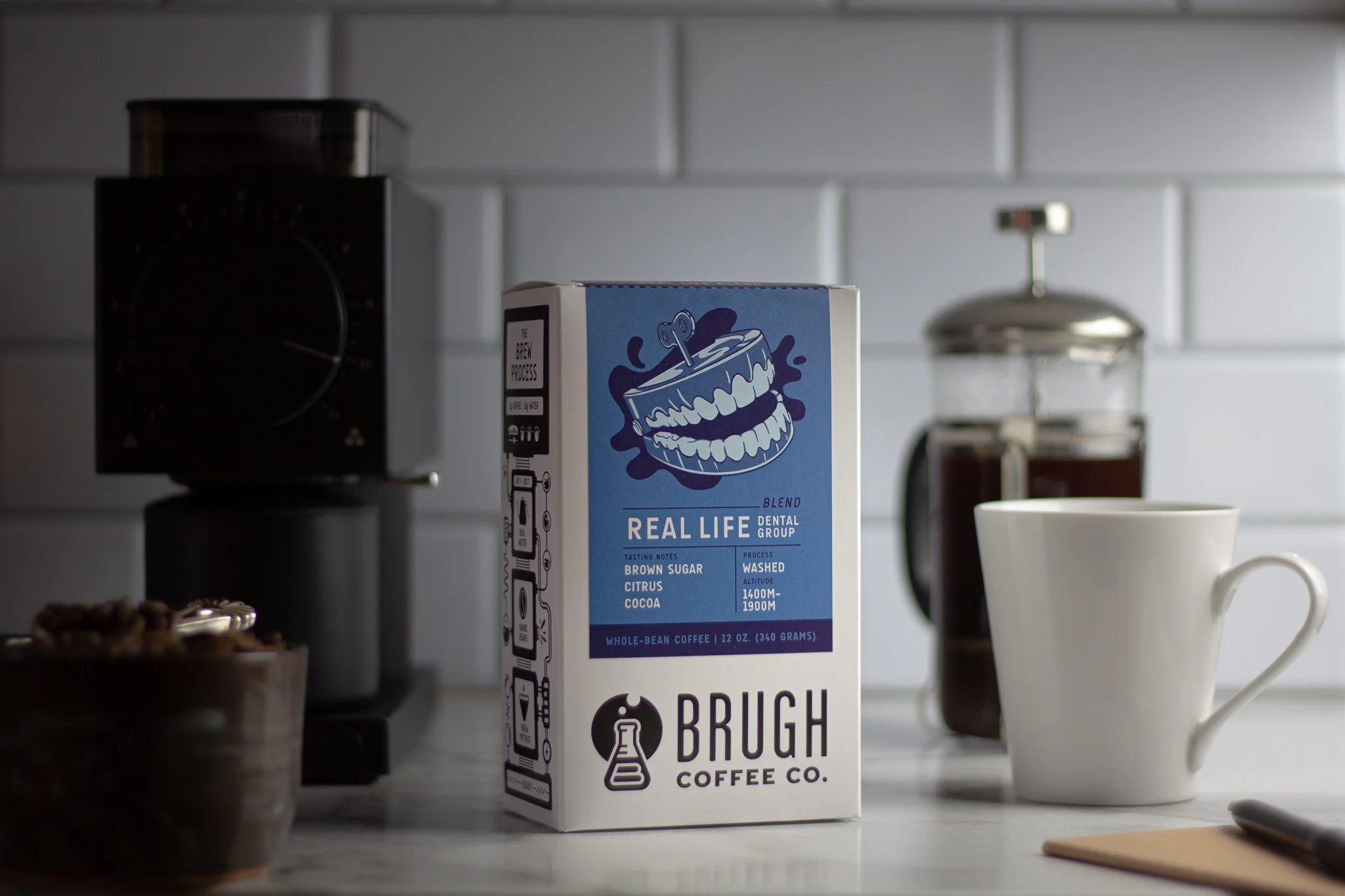 A carton of Brugh Coffee with a blue label featuring a cartoon coffee grinder, placed on a white kitchen counter next to a white mug, a jar of coffee grounds, and a pastry in a bowl, with a coffee grinder in the background.