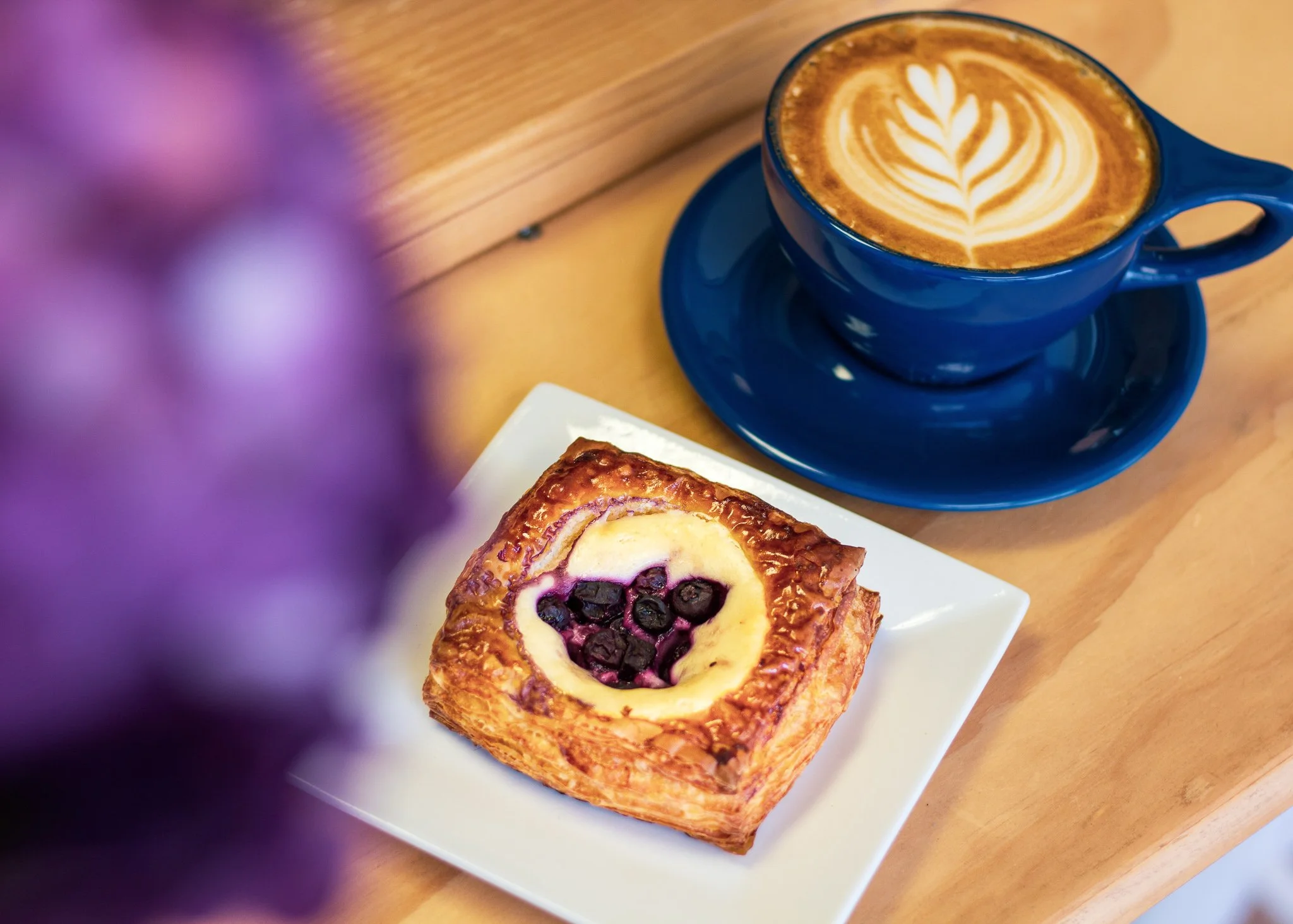 A blue coffee cup with latte art and a square pastry with blueberries on a white plate on a wooden table, with purple flowers blurred in the foreground.