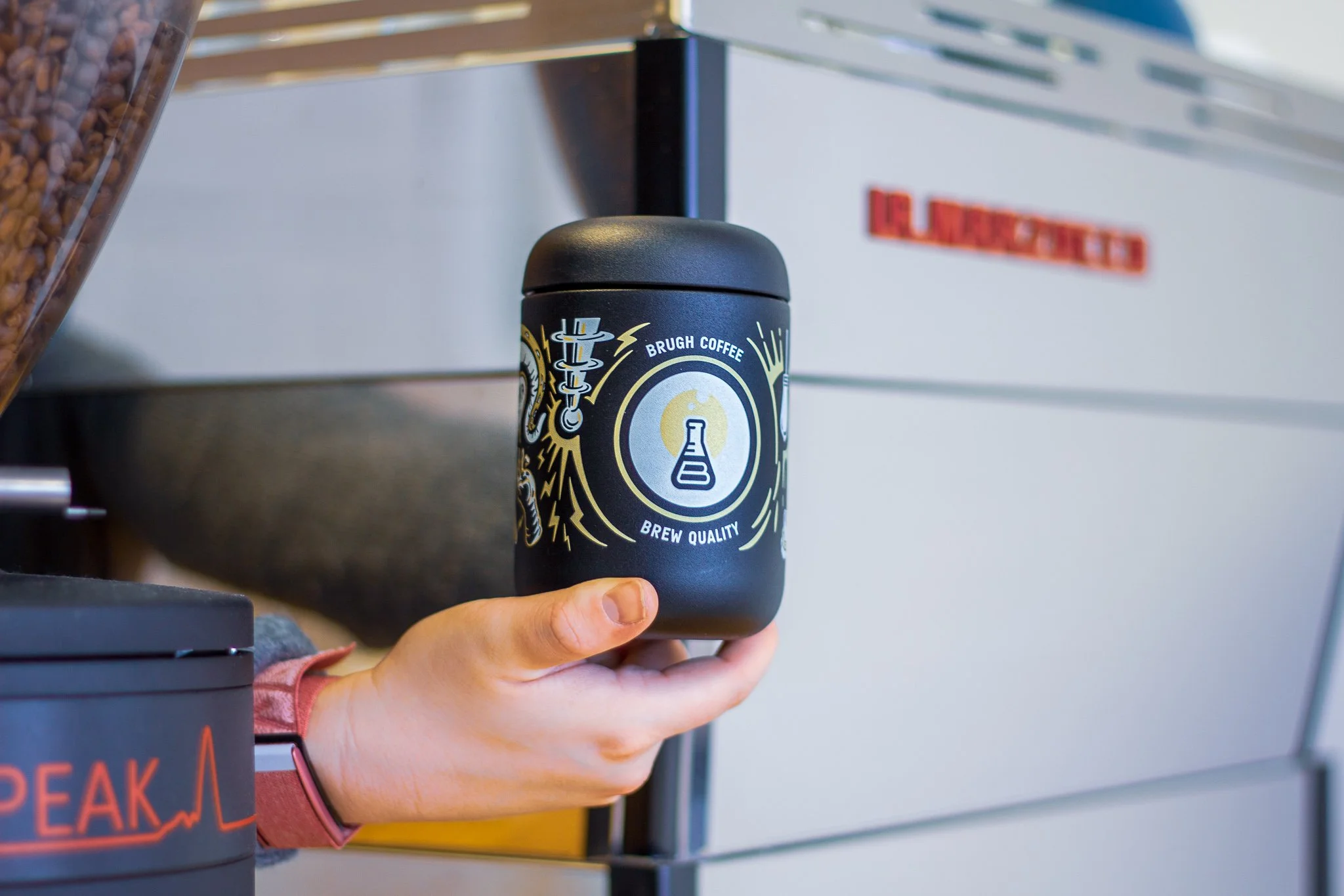 A person holding a black coffee container with a logo featuring a scientific flask, surrounded by lightning and circuit motifs, on a coffee shop counter.