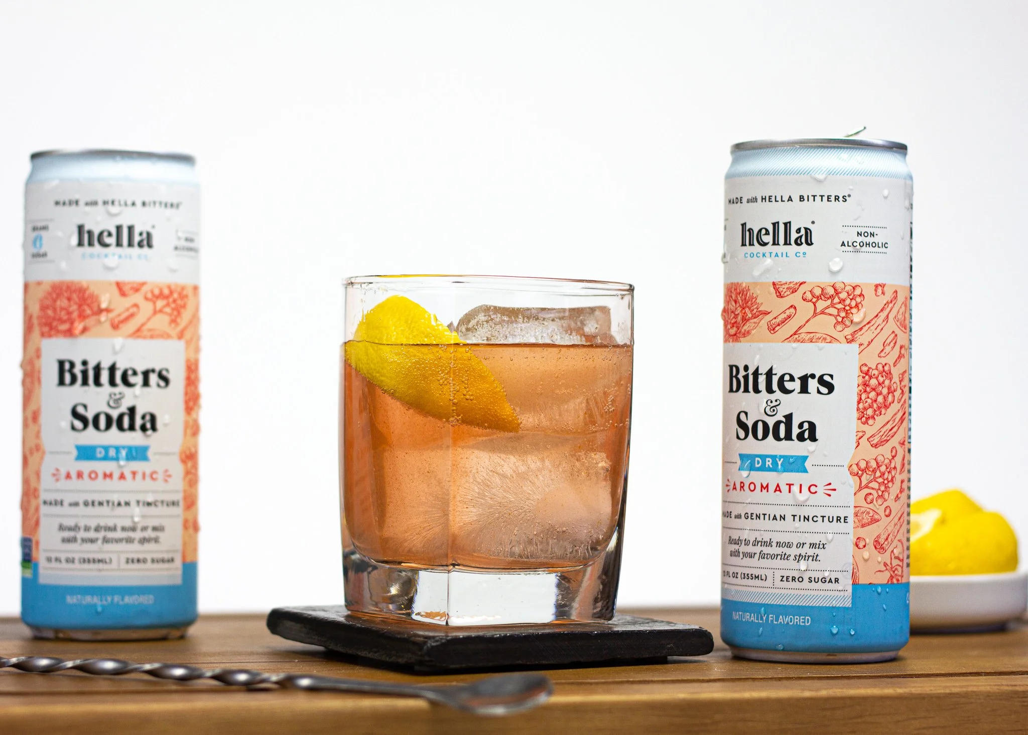 A glass of pink cocktail with a lemon wedge on ice, flanked by two cans of Hella Bitters & Soda, on a wooden surface with a lemon in a small white dish in the background.