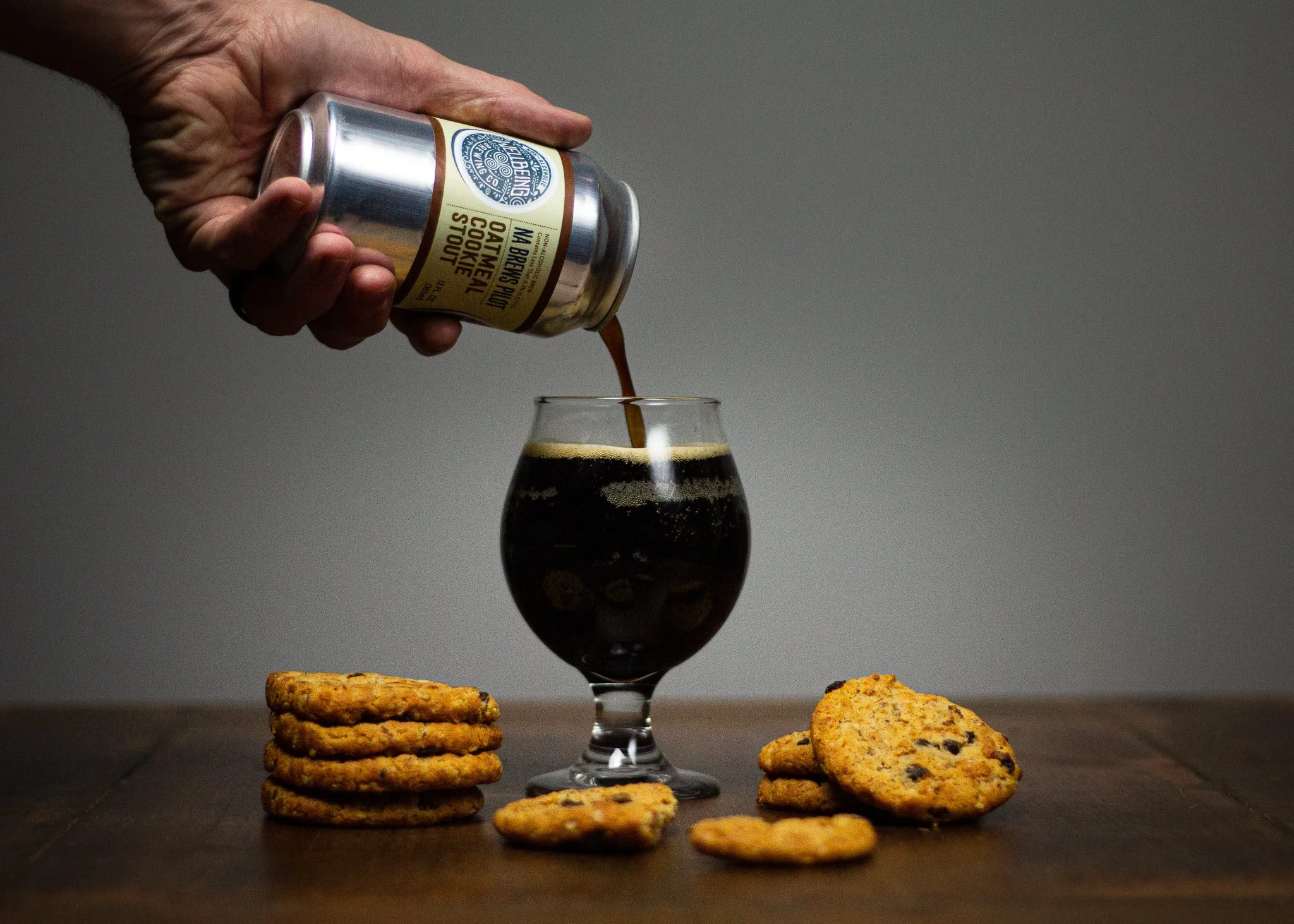 A hand pouring dark cola from a can into a glass with ice, surrounded by cookies on a dark wooden table with a plain grey background.