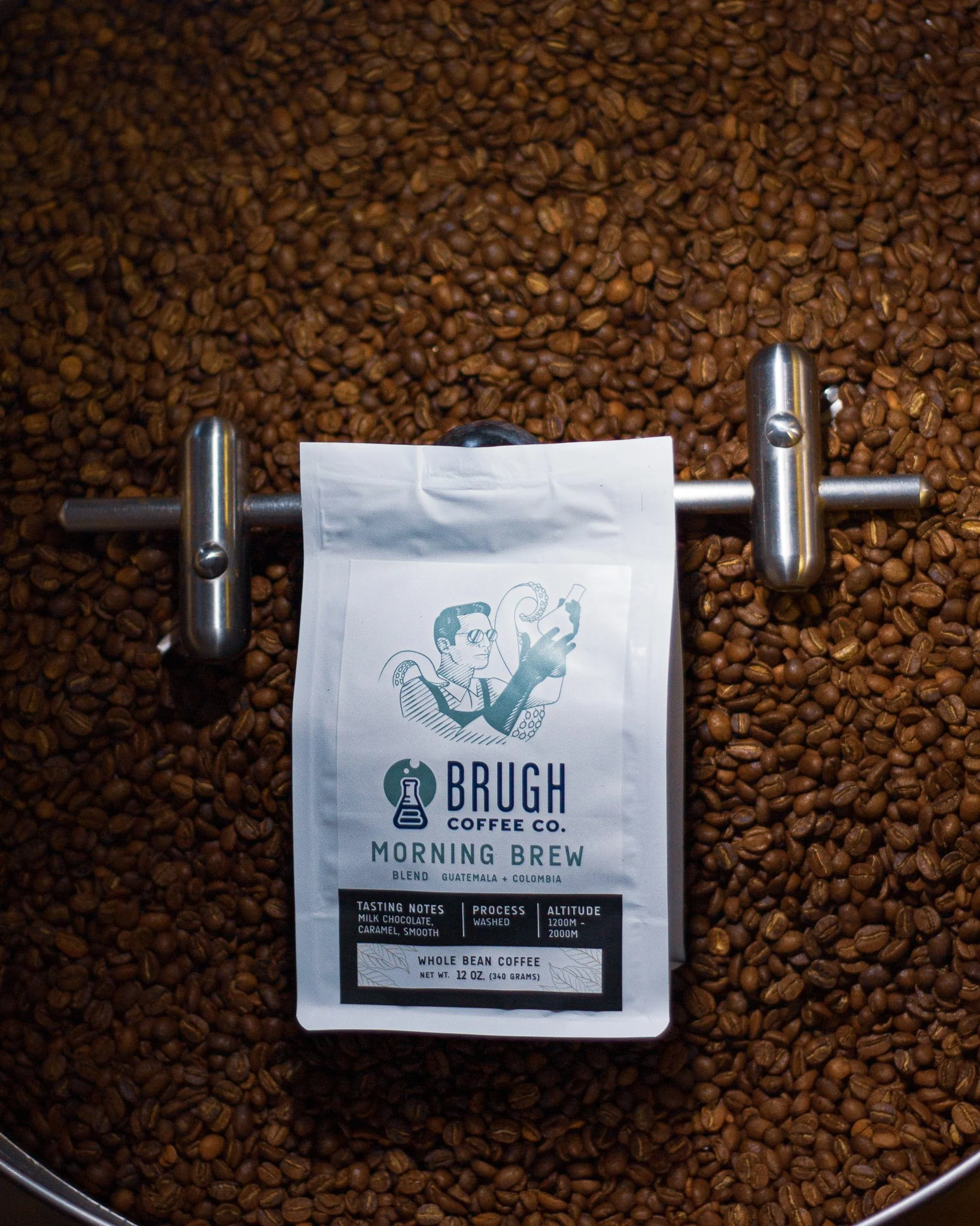 A bag of Brugh Coffee Co. Morning Brew whole bean coffee placed on a bed of roasted coffee beans inside a metal container.