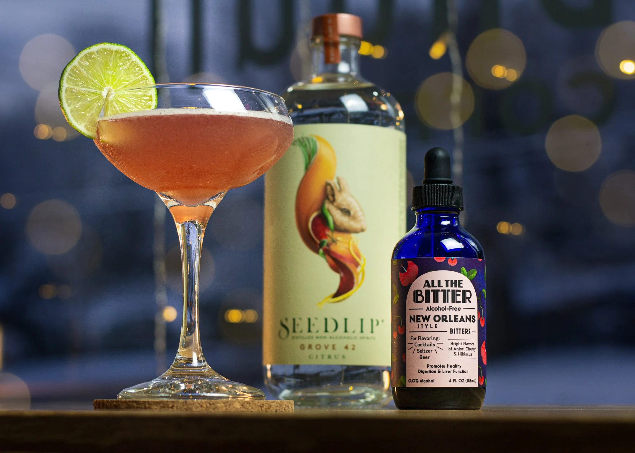 A cocktail with a lime slice garnish on a stemmed glass, a bottle with colorful label featuring a hare and fruit, and a small dark blue bottle labeled "All The Bitter" with flavors of cherry, hibiscus, and bitter, set against a blurred background.