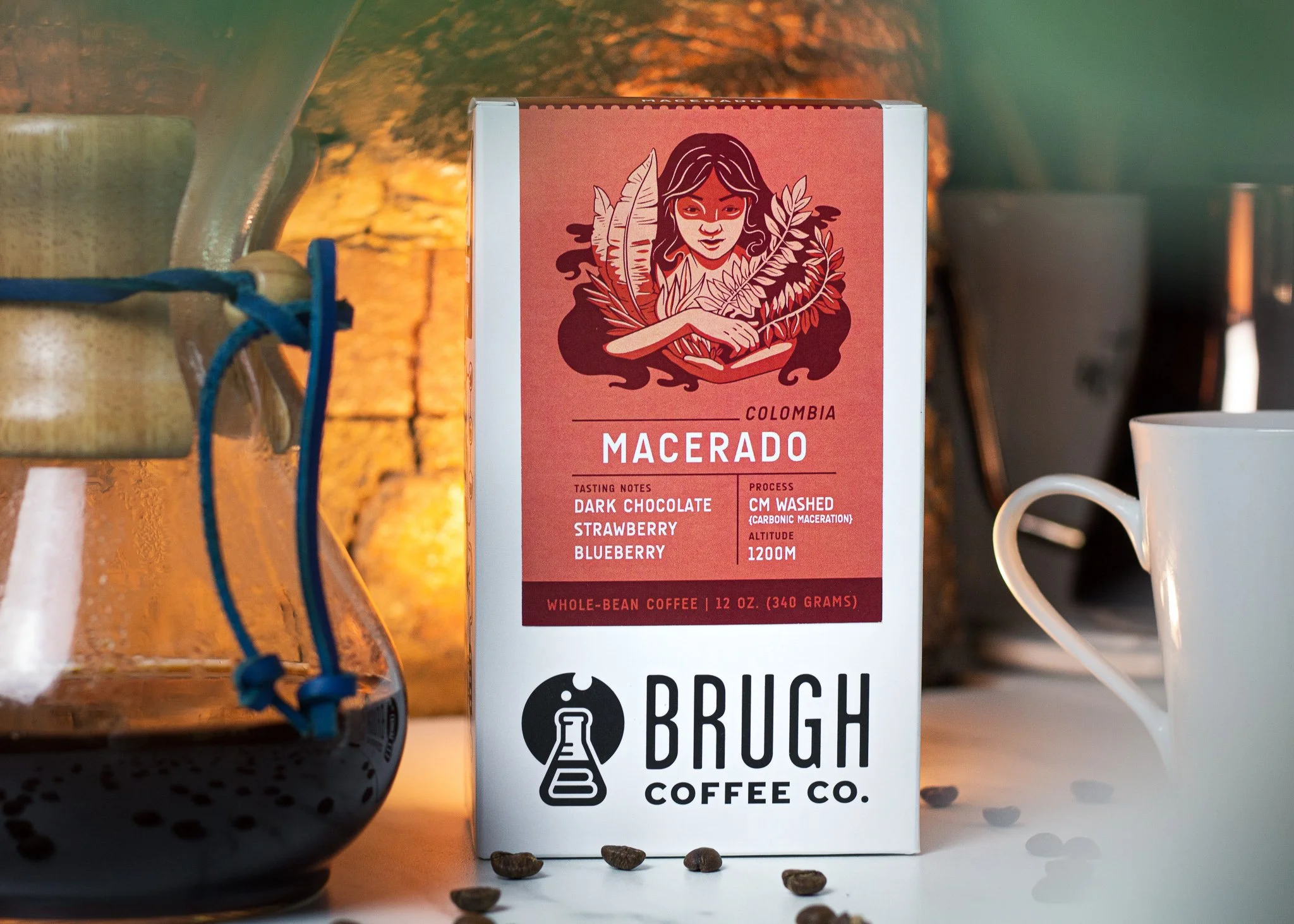 A box of Brugh Coffee Macorado coffee from Colombia, featuring a stylized illustration of a woman with long dark hair holding plants, with brewing process details and taste notes of dark chocolate, strawberry, and blueberry, placed on a table surroun