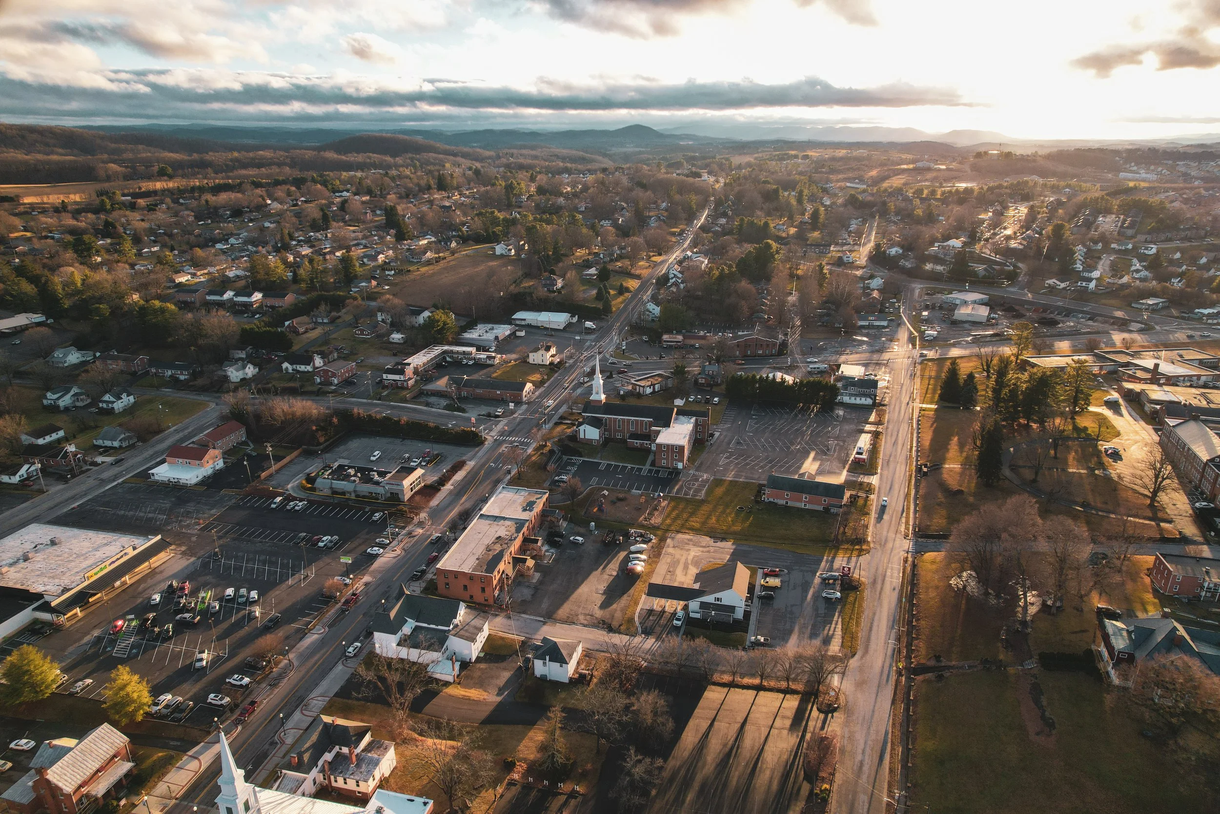 An aerial view of a small town with commercial and residential buildings, parking lots, trees, and roads during sunset.