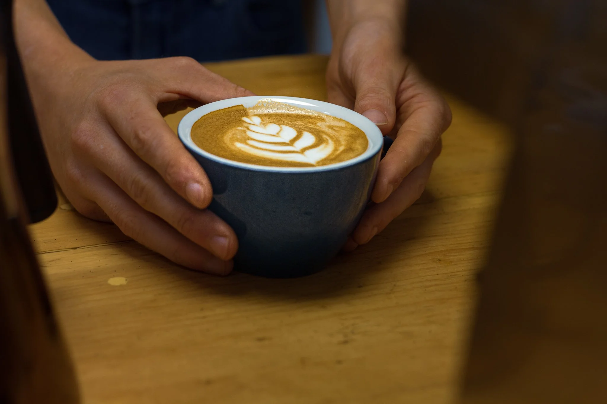 Person holding a blue mug of coffee with latte art, sitting at a wooden table.