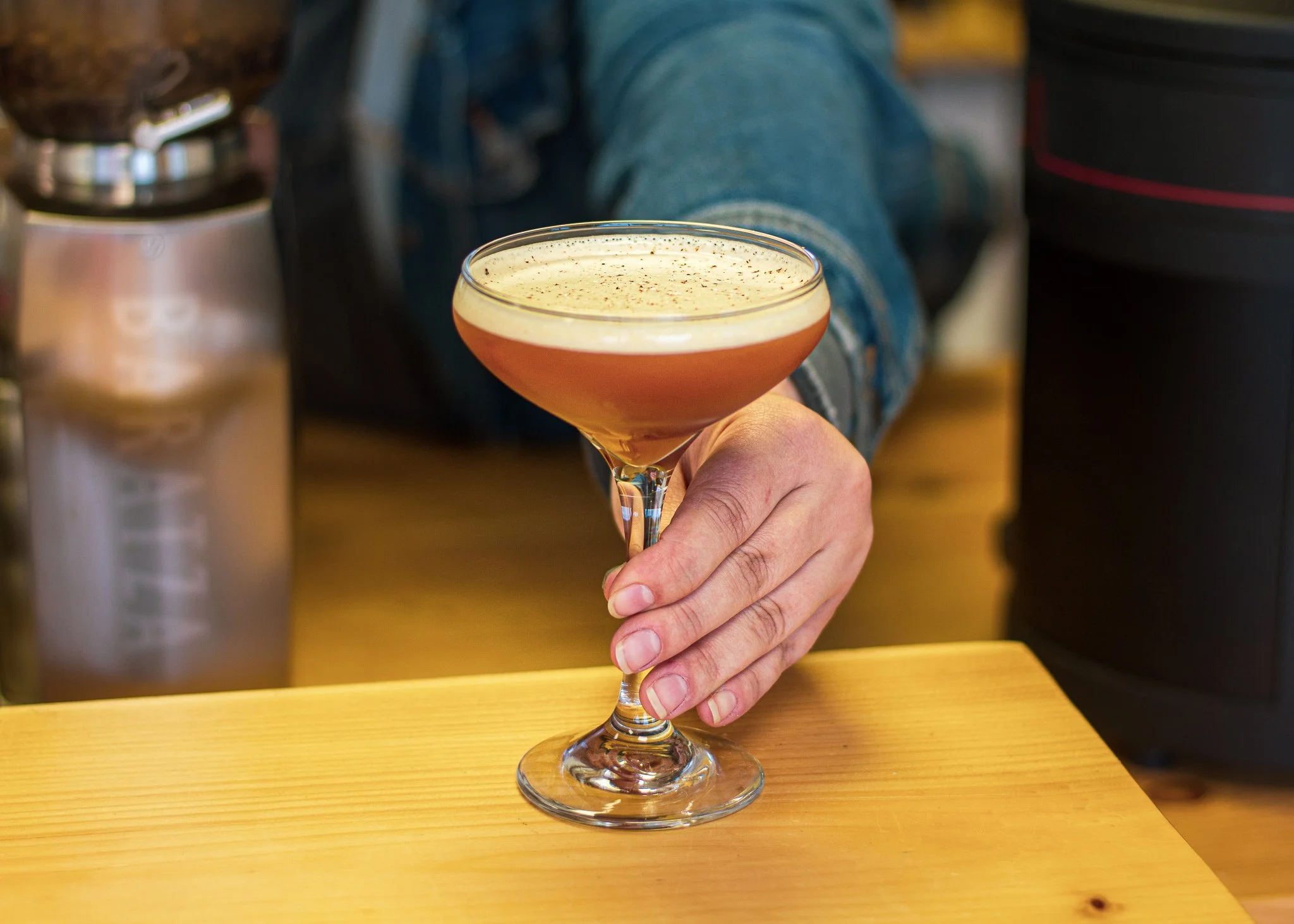 A hand holding a cocktail glass with a frothy, pinkish drink garnished with black specks, on a wooden surface.