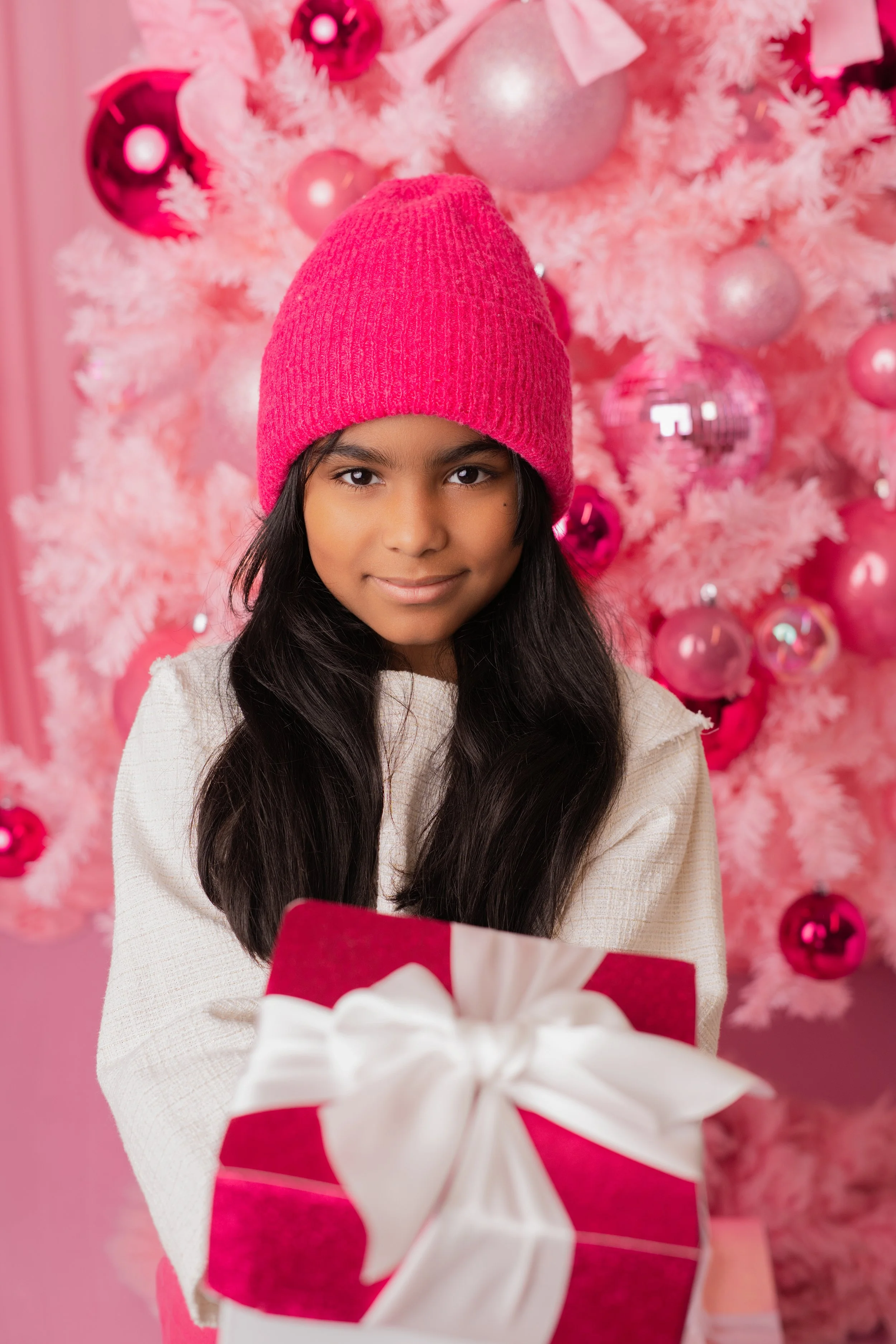 A young girl with long black hair wearing a pink knit hat and a white sweater, holding a red gift box with a white ribbon, standing in front of a pink Christmas tree decorated with pink and silver ornaments.