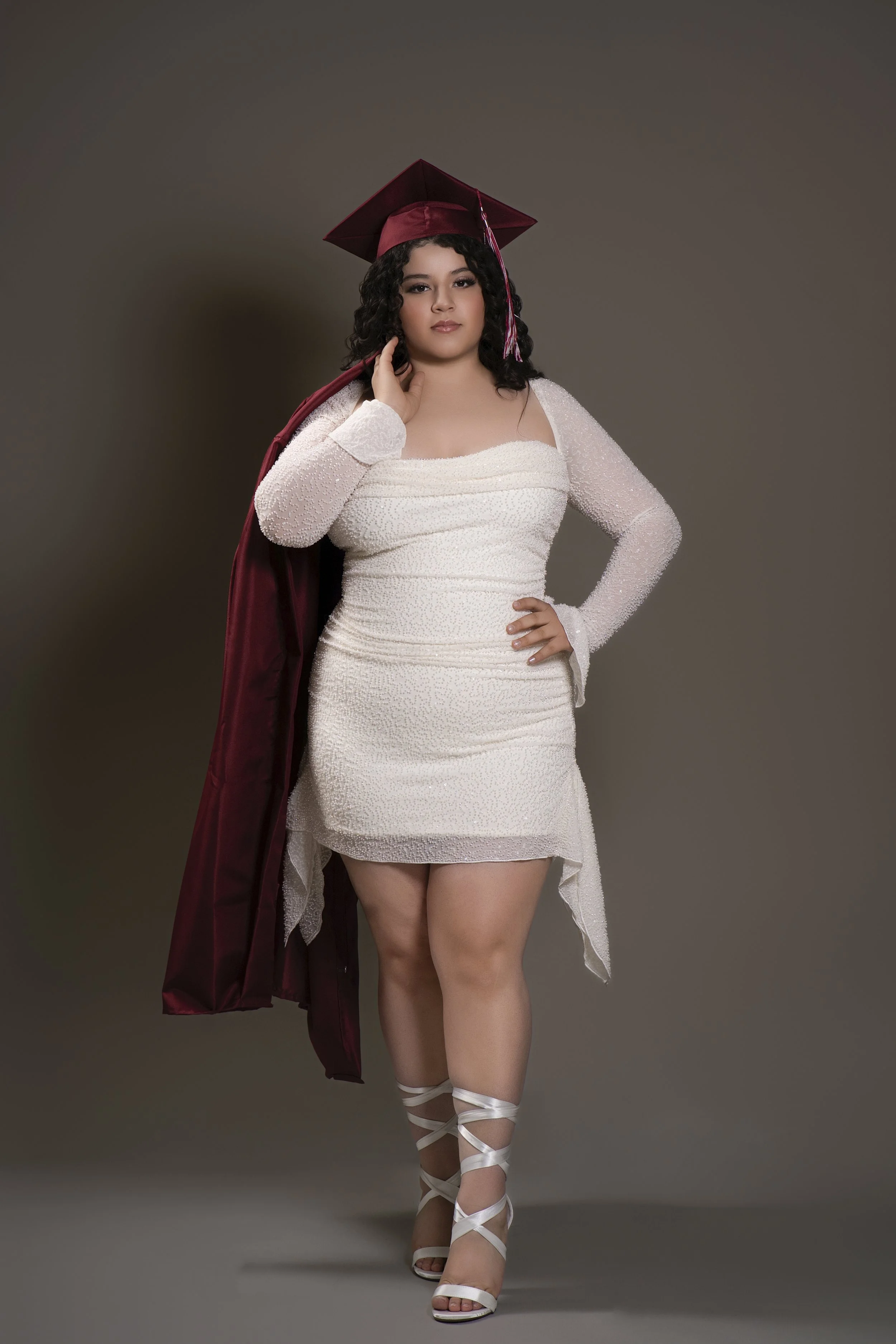 Woman in maroon graduation cap and gown with long white dress and white strappy sandals.