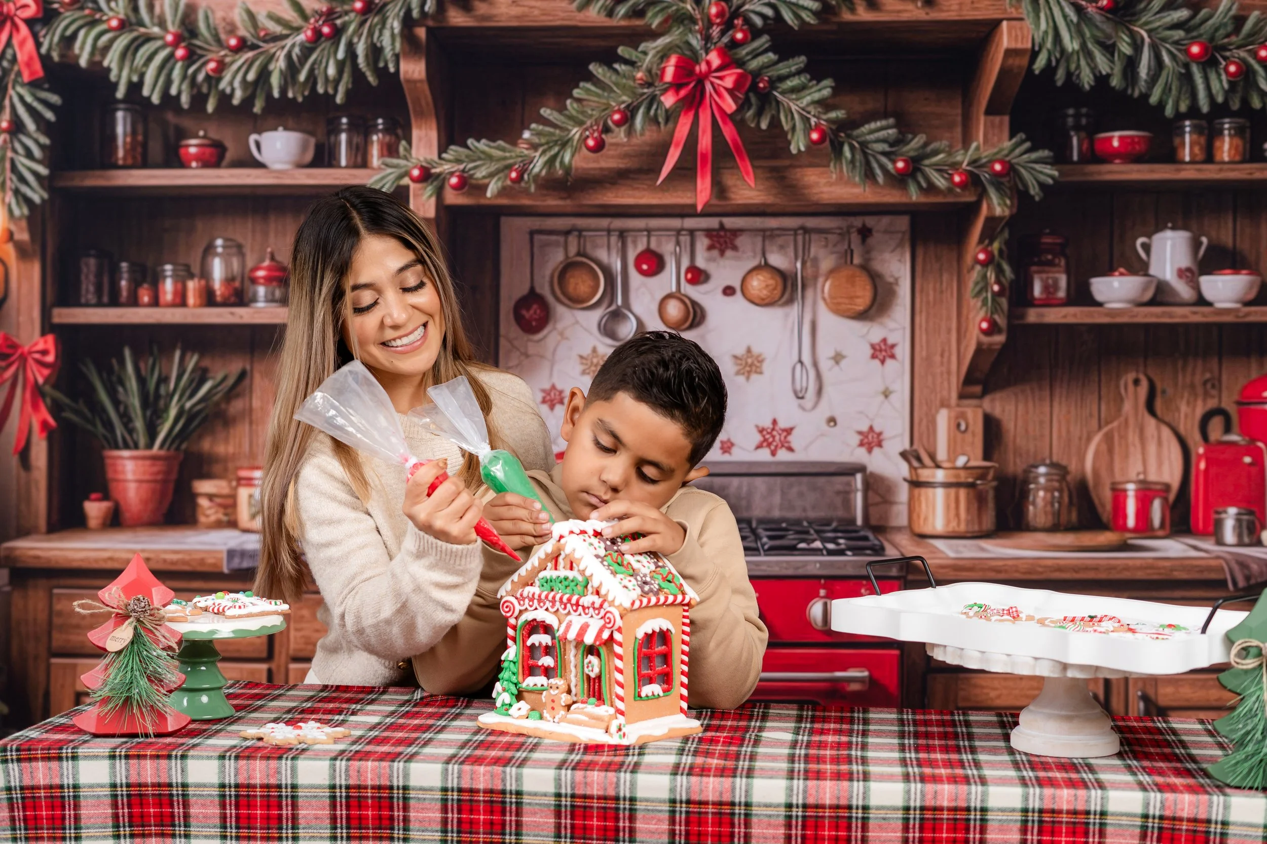 A woman and a young boy decorating a gingerbread house at a Christmas-themed kitchen table. The woman is holding a piping bag and smiling, while the boy is concentrating on placing decorations. The table has a red and green plaid tablecloth with Chri