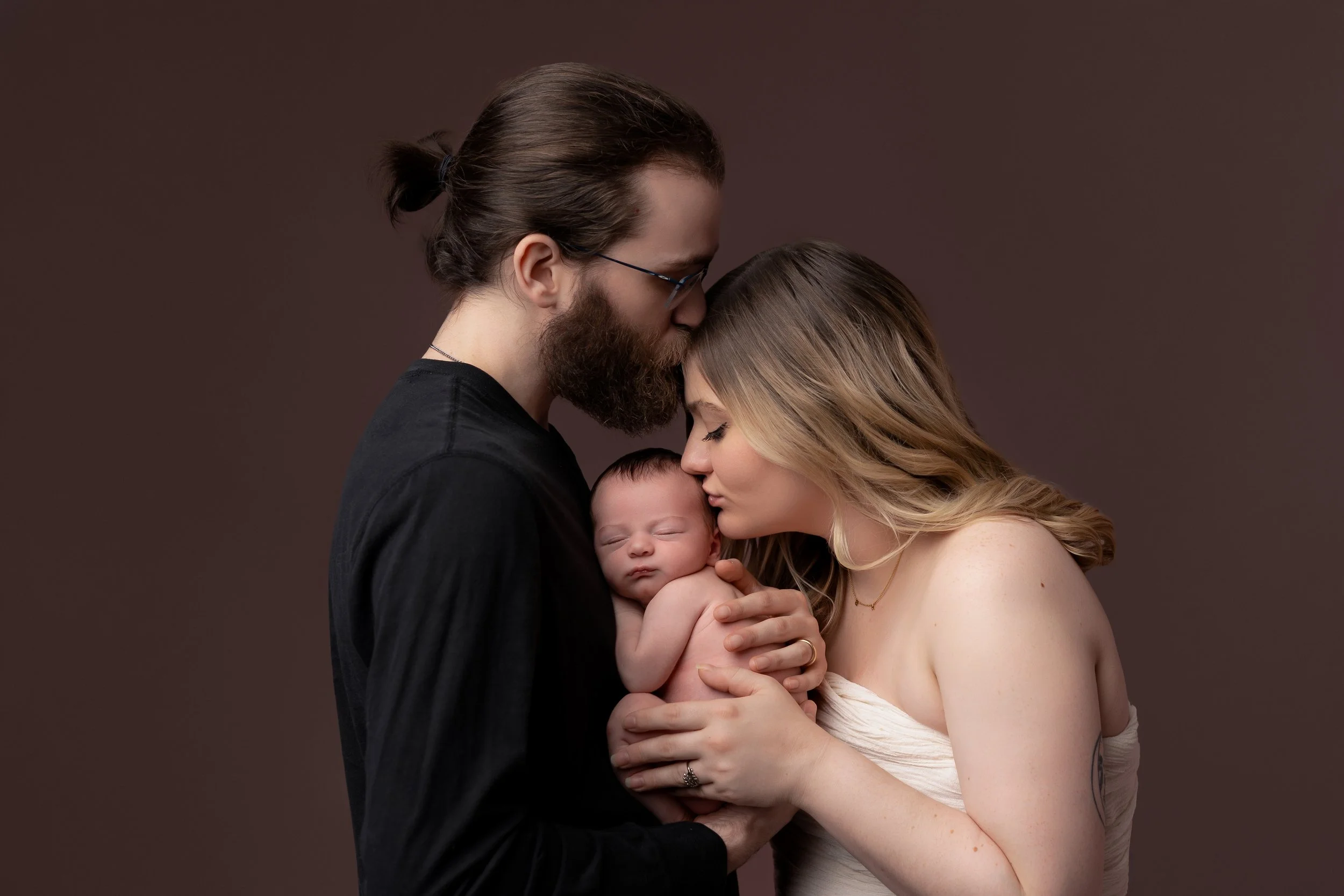A couple lovingly holding a newborn baby. The man kisses the baby's head, while the woman gently cuddles the baby. The background is plain and neutral.