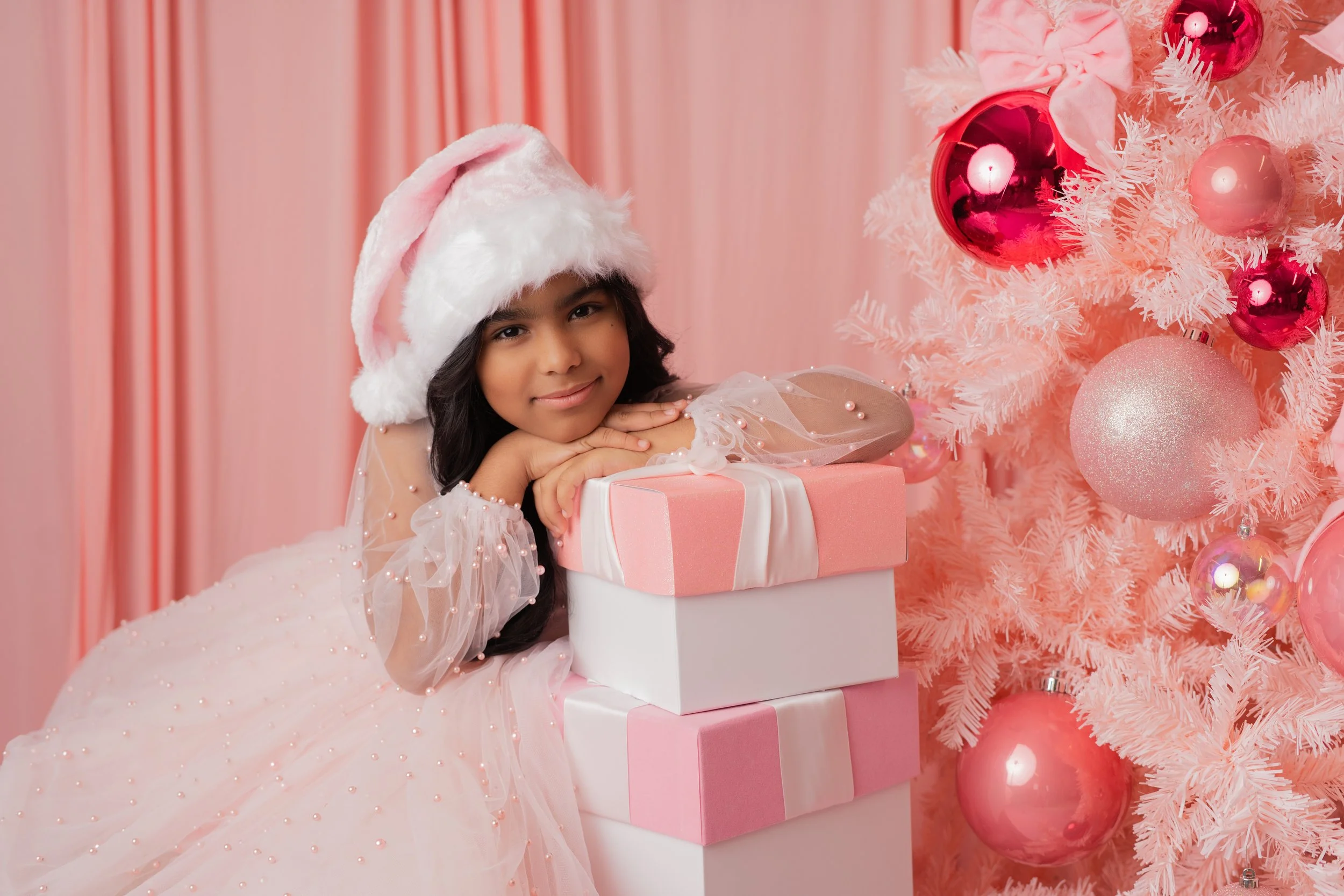 A young girl in a pink dress and Santa hat leaning on stacked pink and white gift boxes, smiling near a pink Christmas tree with pink and rose ornaments.