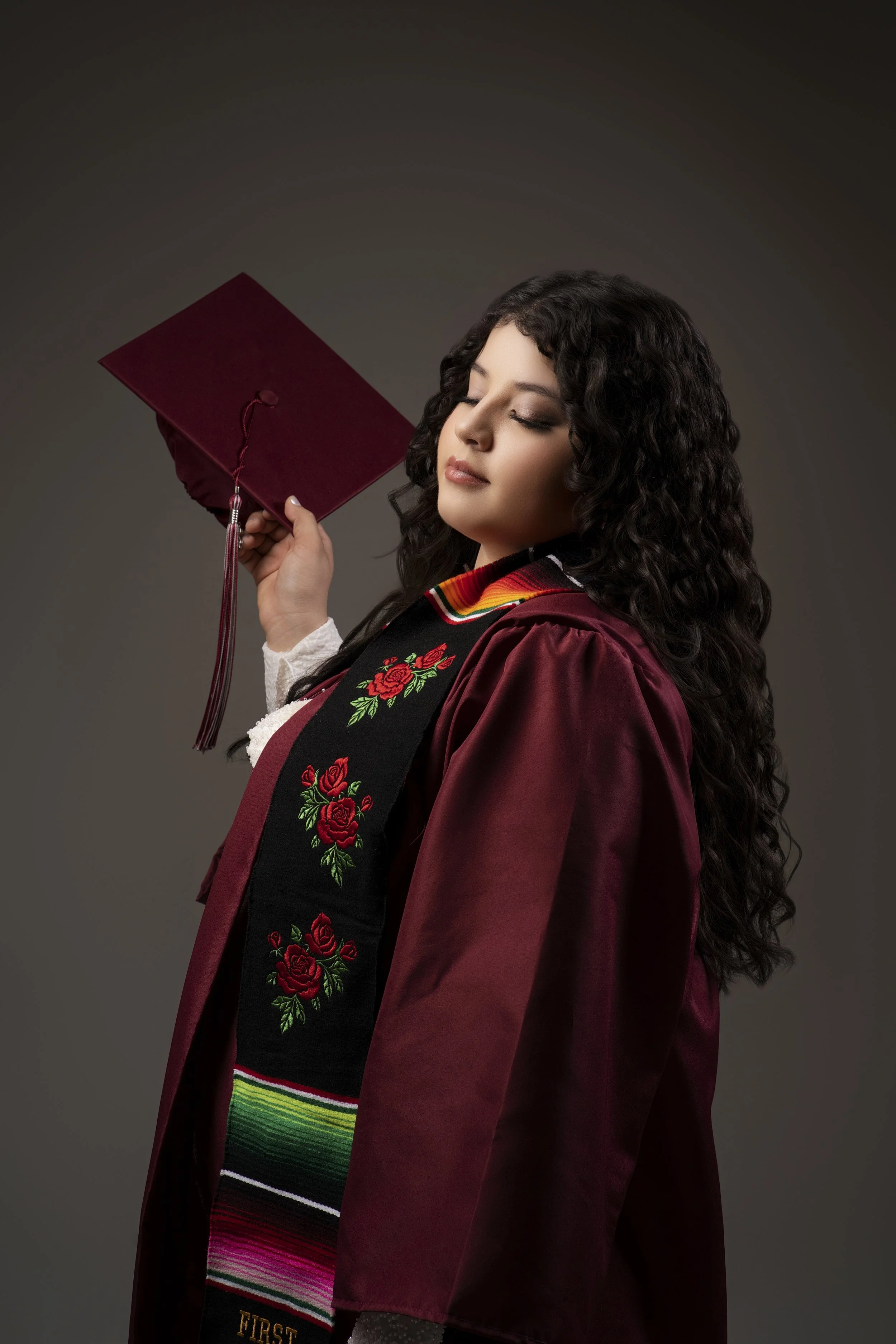 A graduate in a maroon cap and gown holding a mortarboard. She wears a stole with rose embroidery and colorful stripes.