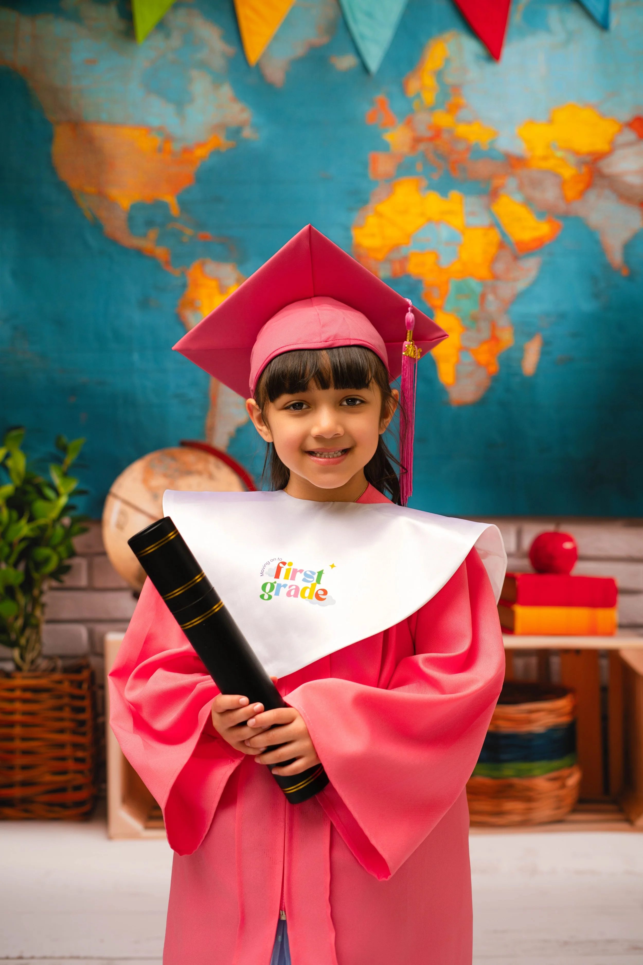Young girl dressed in a pink graduate gown and cap, holding a diploma, standing in front of a world map classroom decoration.