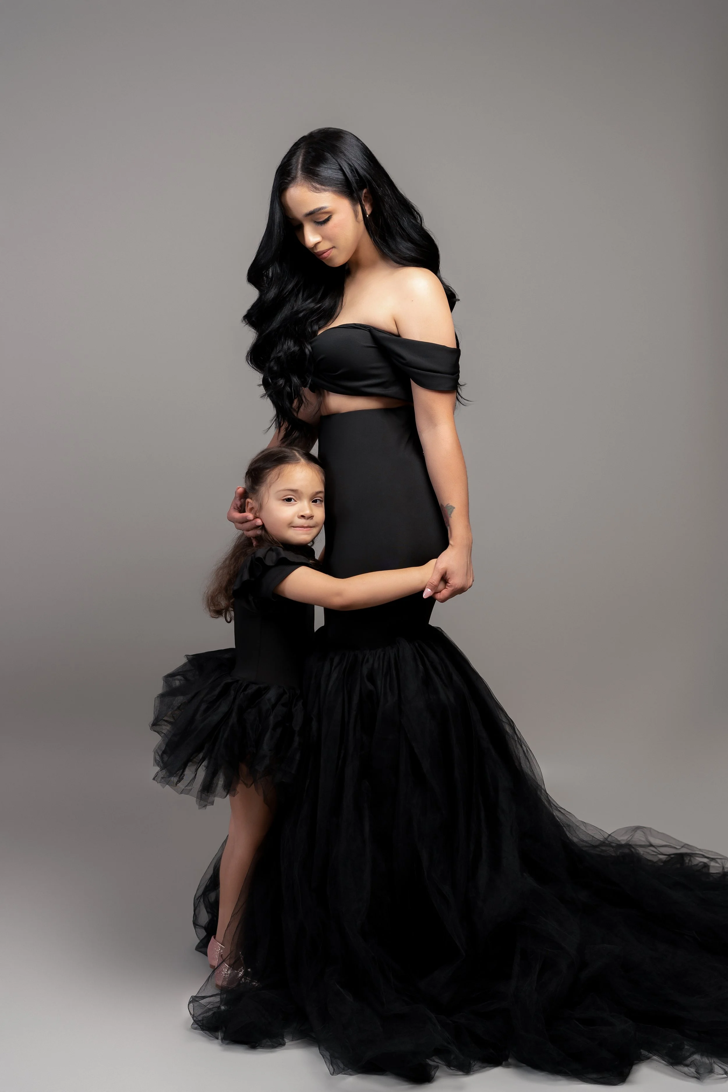 A woman in an elegant black gown standing with a young girl in a matching black dress, both looking down and embracing each other against a gray background.