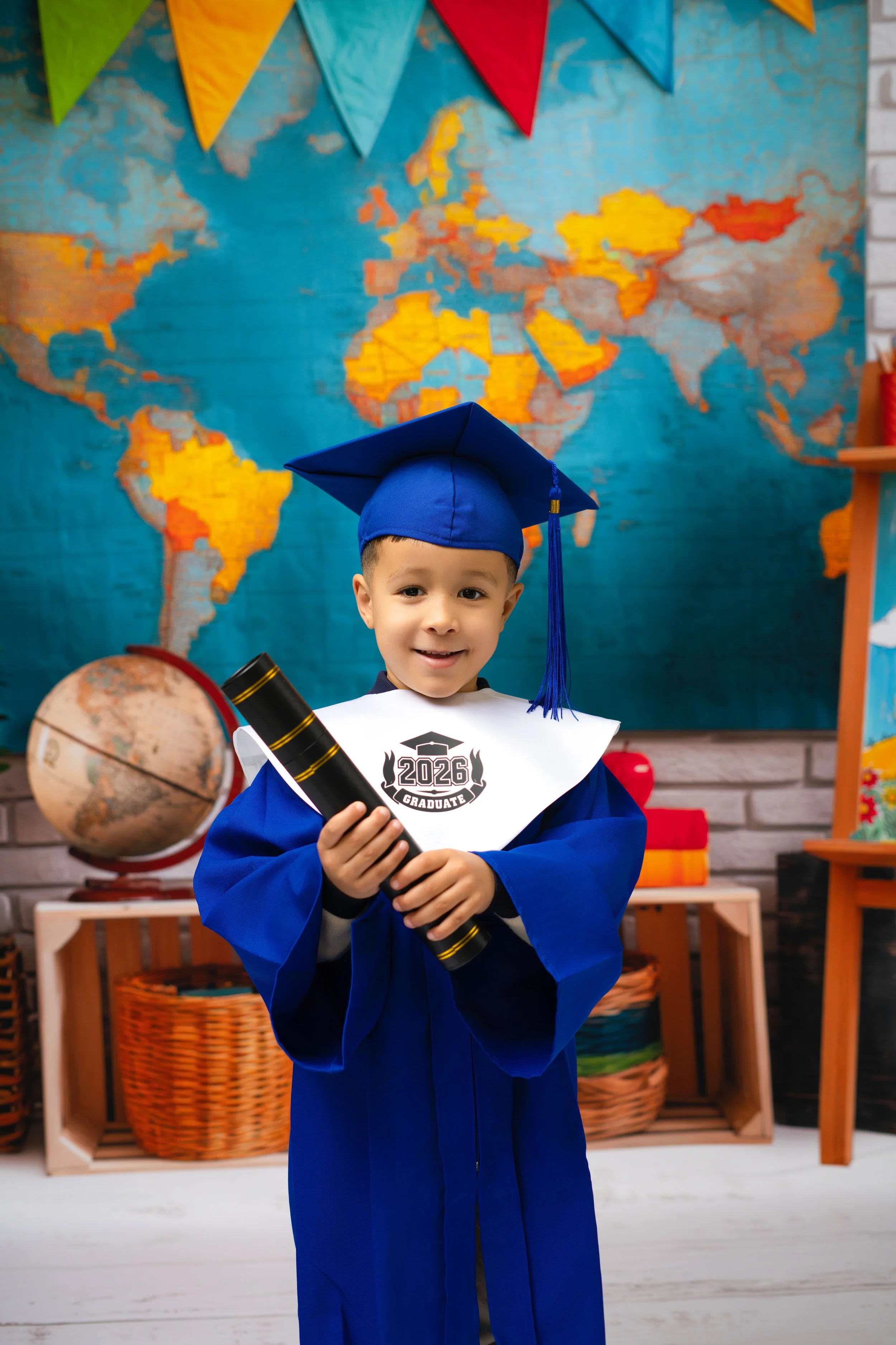 Young boy in blue graduation cap and gown holding a diploma in classroom with world map and globe in background.