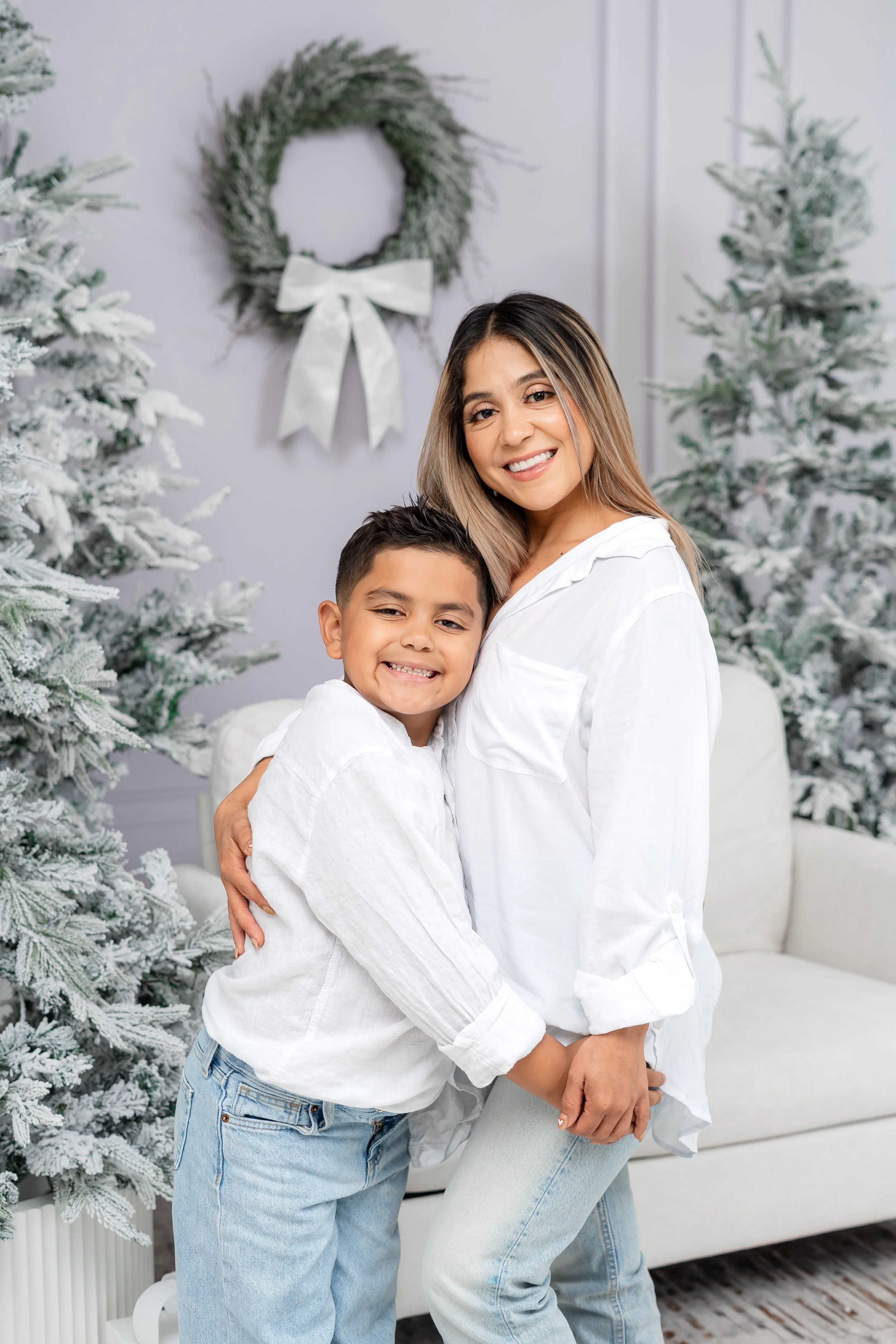 A woman and a boy hugging and smiling, with snow-dusted Christmas trees and a wreath with a bow in the background.