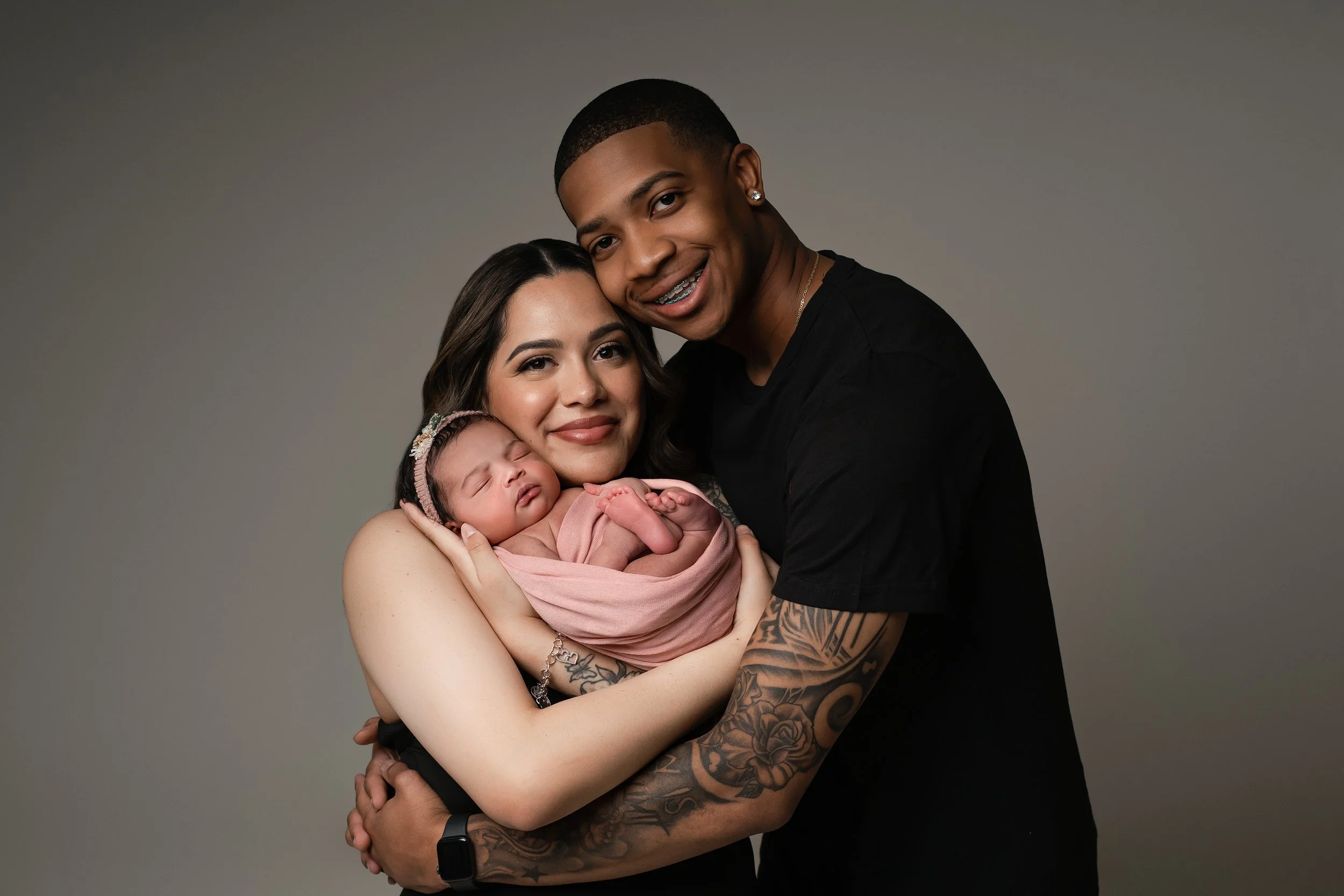 A couple smiling while holding a sleeping newborn wrapped in a pink blanket, against a plain background.