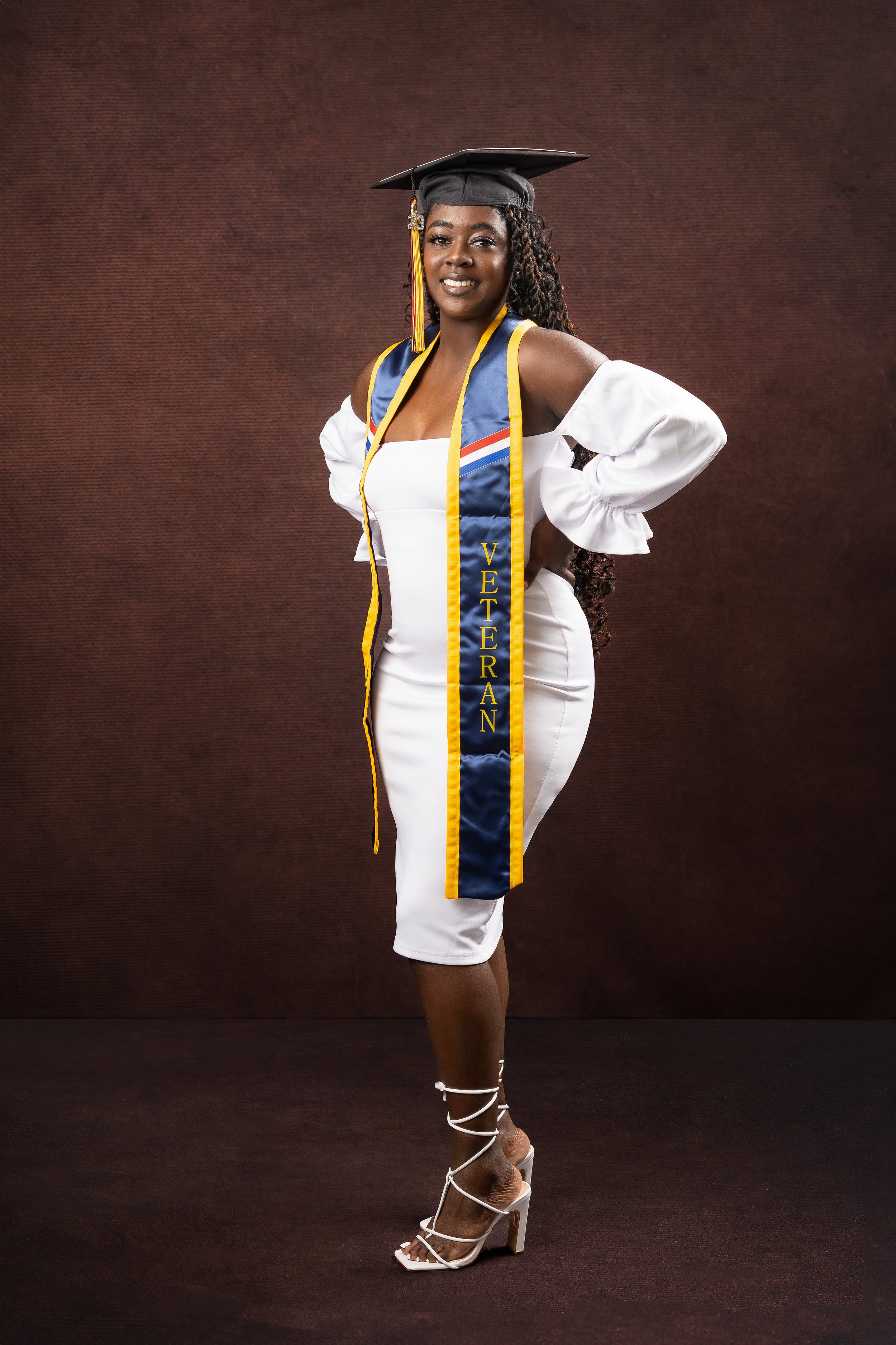 A graduate poses wearing a white dress, graduation cap, and a blue sash labeled "Veteran."