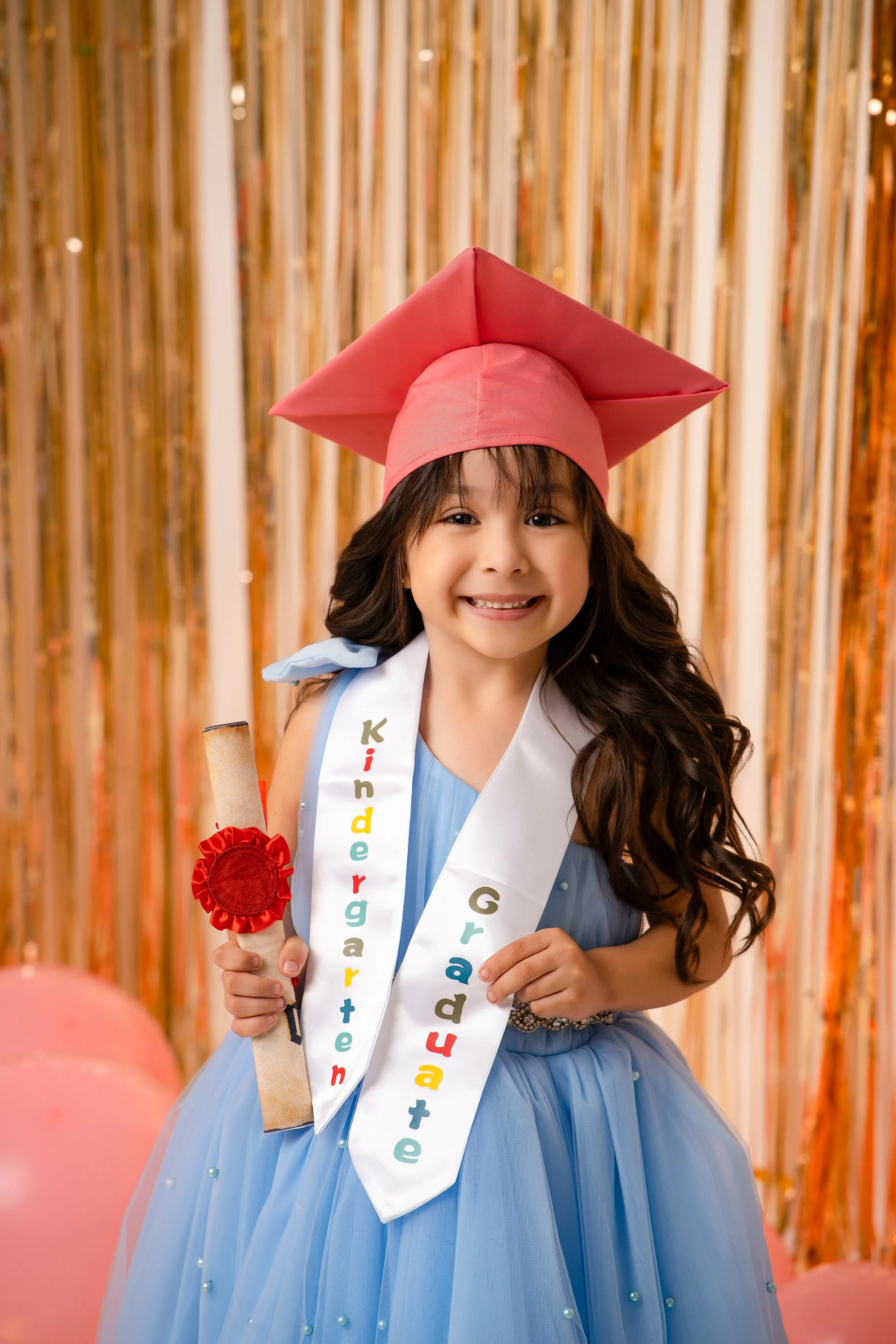 Young child wearing a pink graduation cap and a blue dress with a kindergarten graduate sash, holding a diploma in front of a gold streamer backdrop.