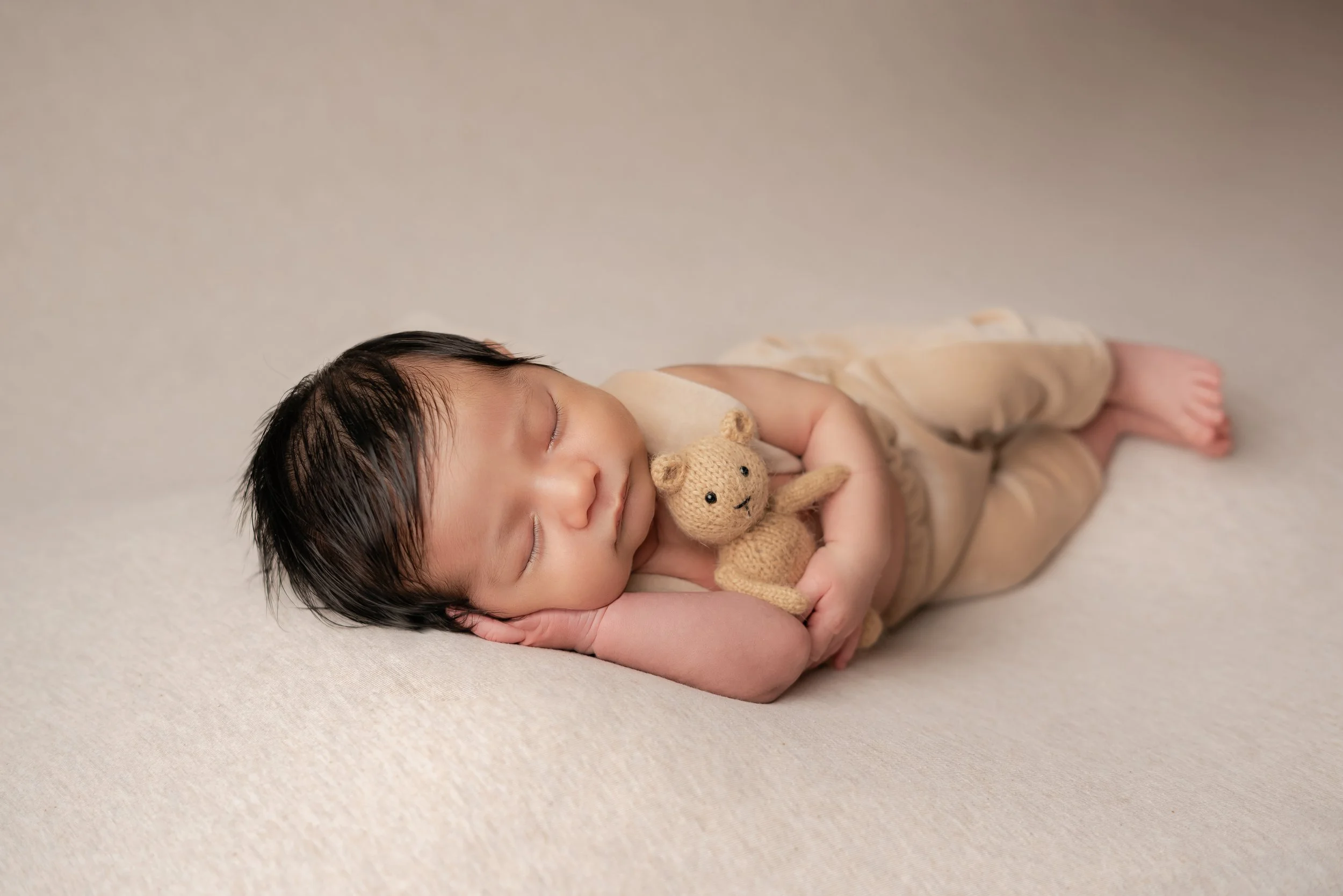 Newborn baby sleeping while holding a small stuffed teddy bear, dressed in neutral-colored clothing, on a soft beige surface.