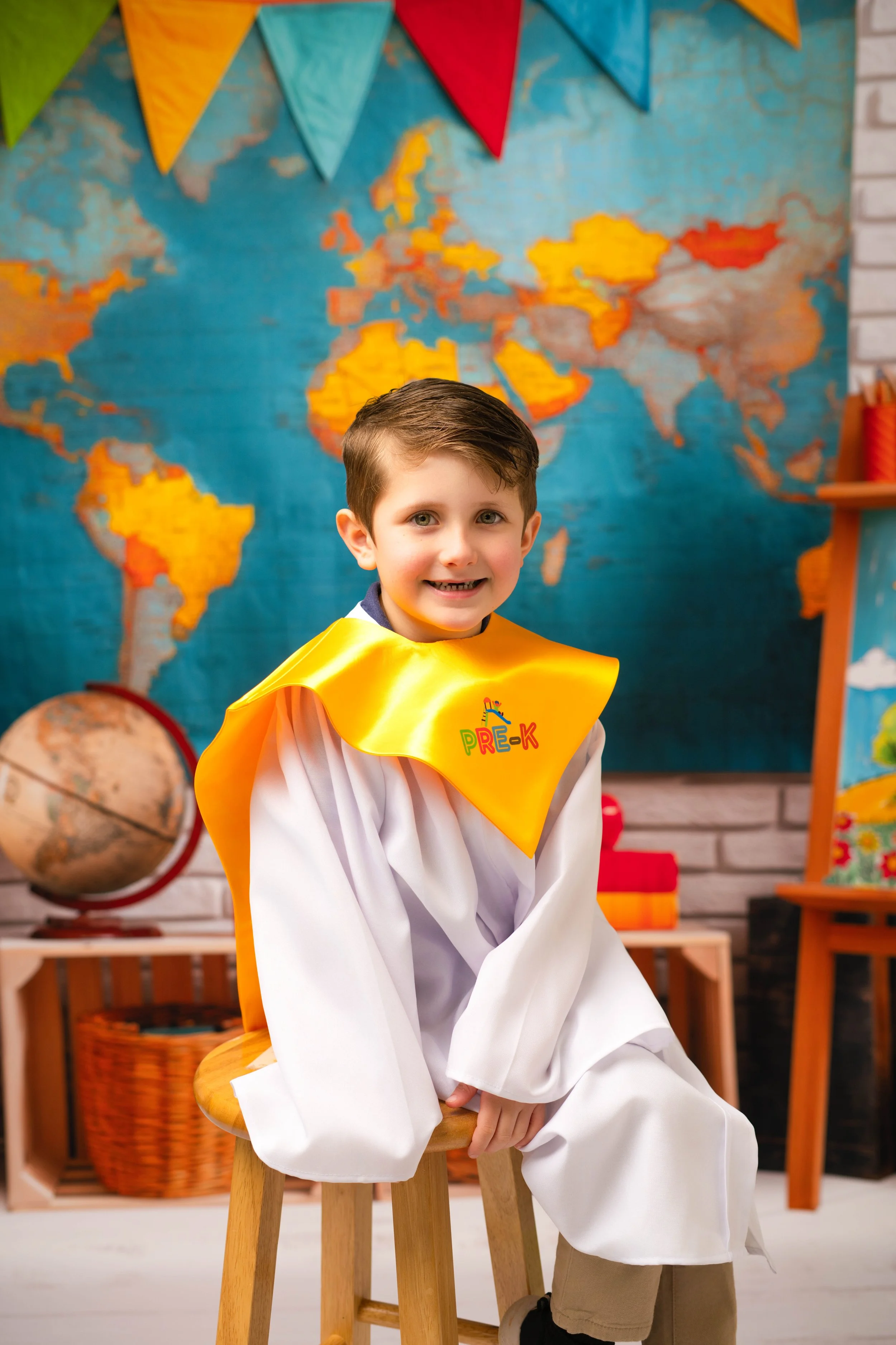 A young boy wearing a white gown and a yellow cape with 'Pre-K' printed on it, sitting on a wooden stool in a classroom with a world map, globe, and educational decorations in the background.