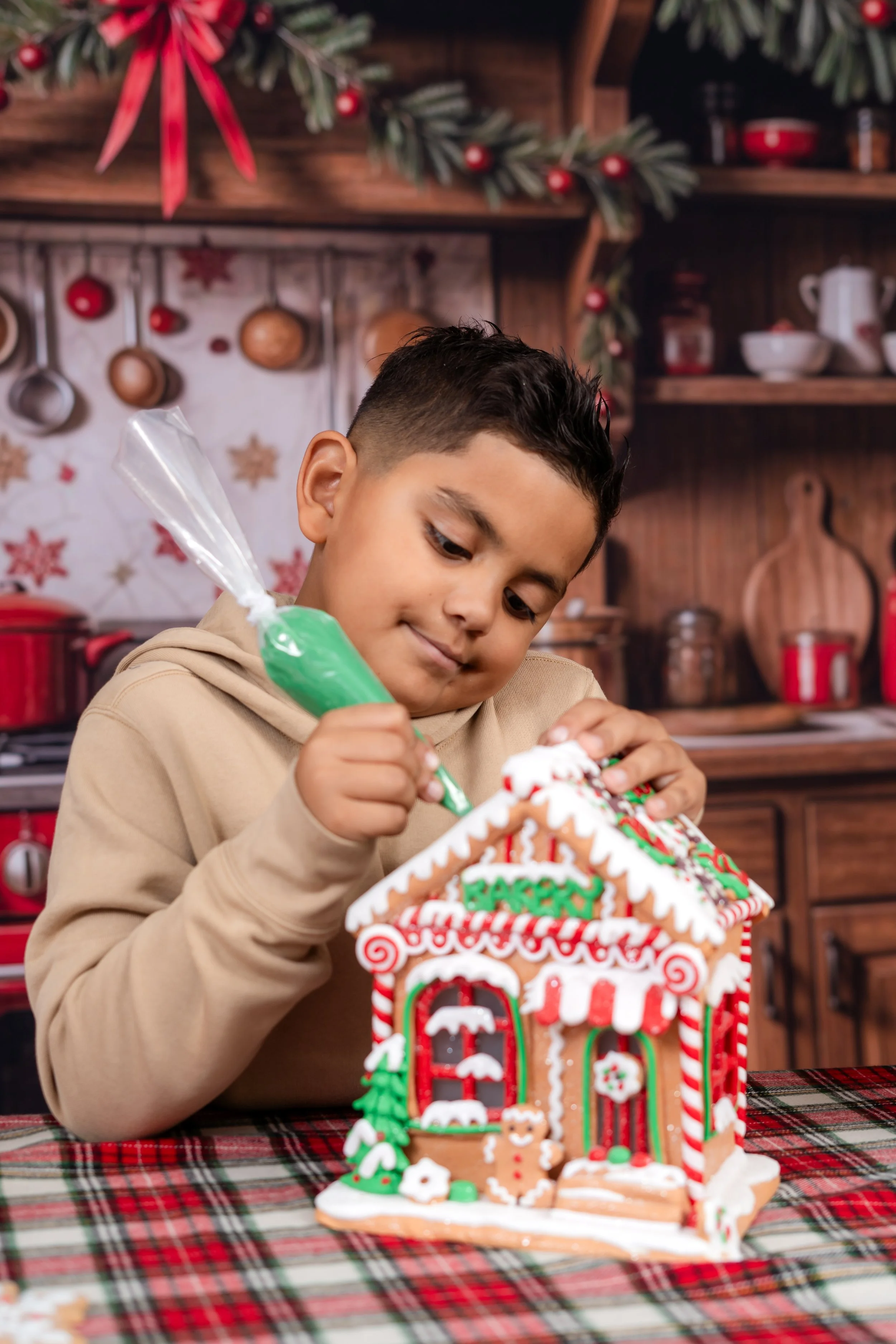 A young boy decorating a Christmas gingerbread house with green icing in a festive kitchen.