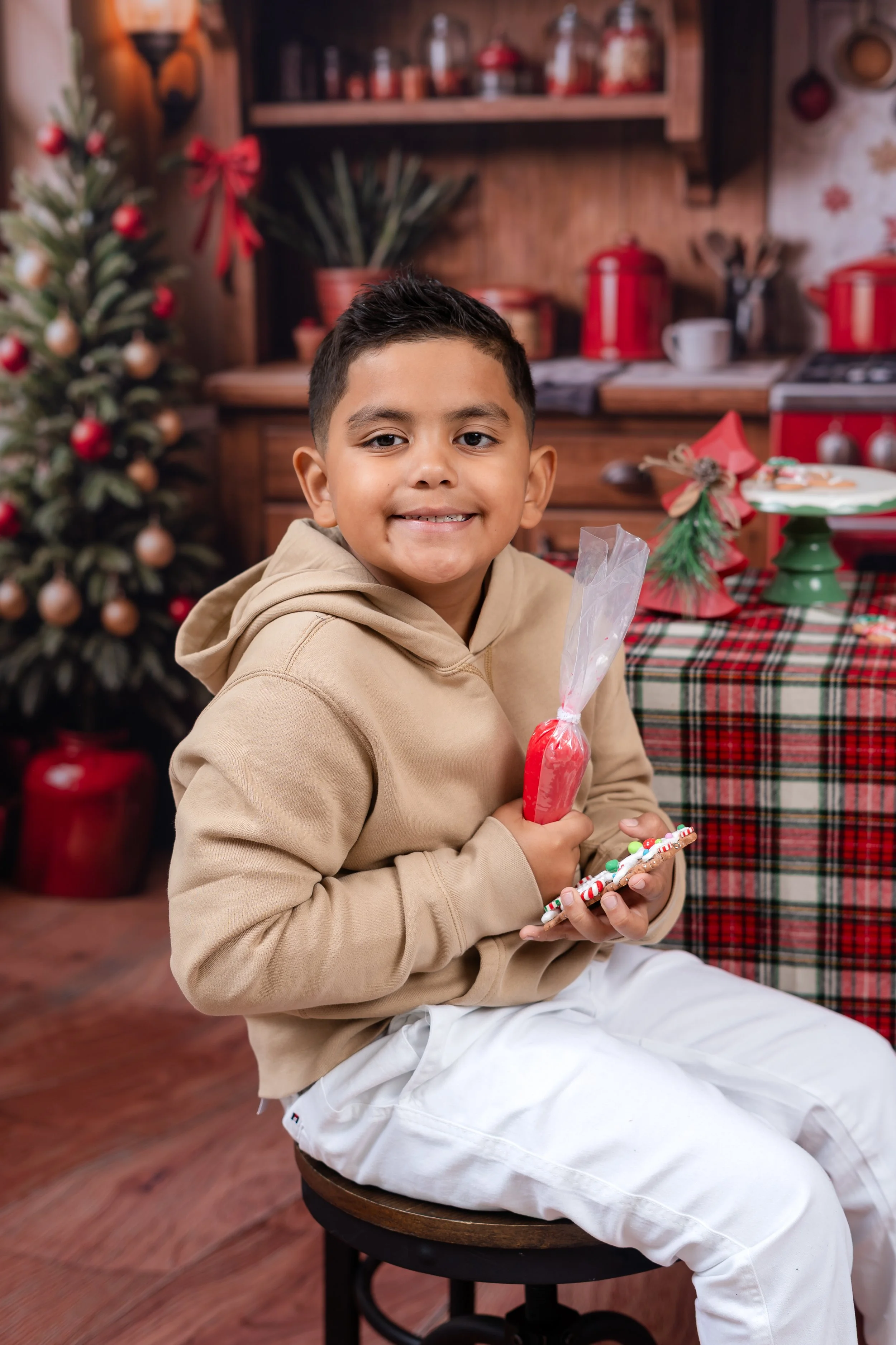 A young boy sitting on a stool, smiling and holding holiday treats in a festive Christmas setting with a decorated tree and wrapped presents.