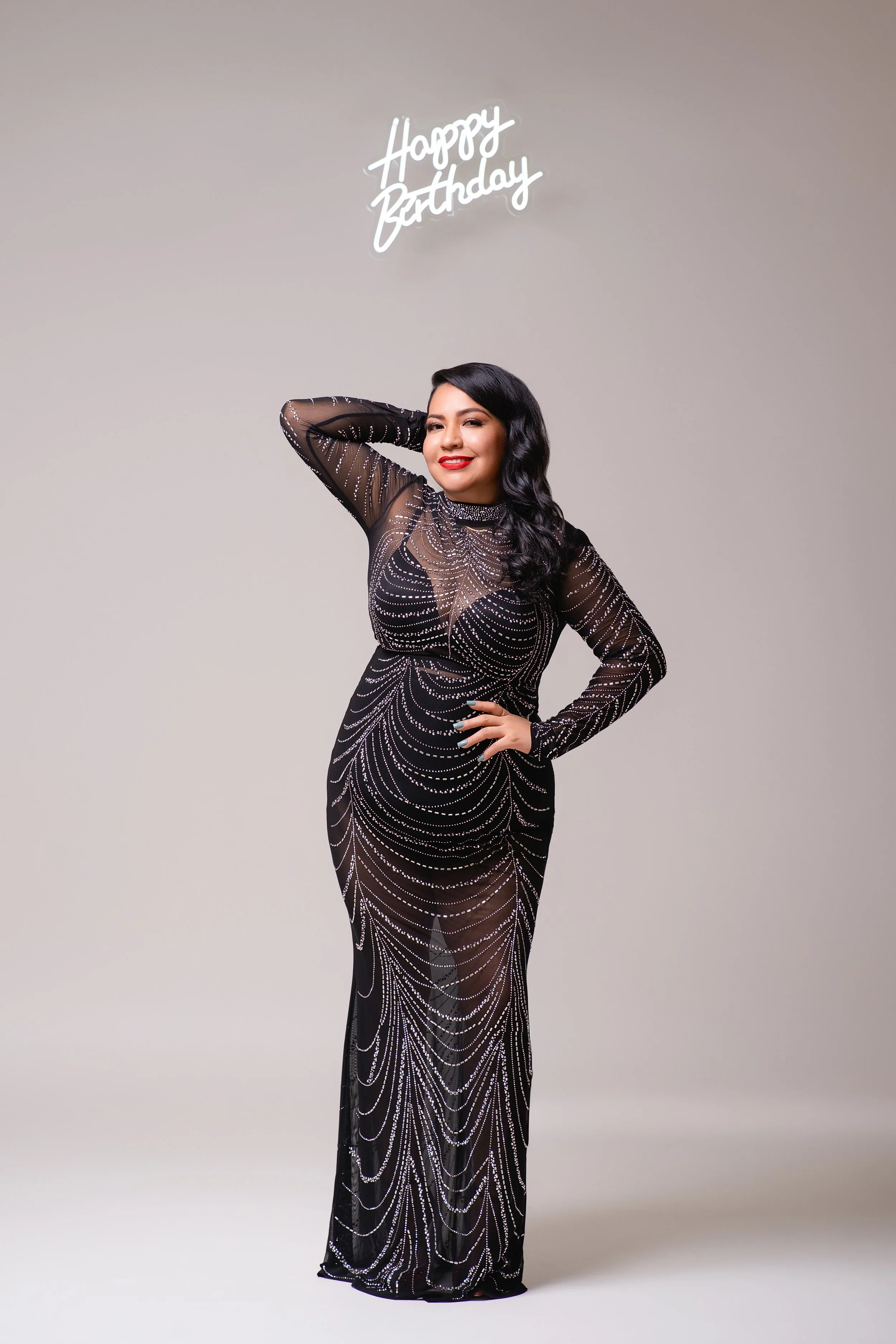 Person in a black sequin gown, posing with one hand on hip, under "Happy Birthday" sign.