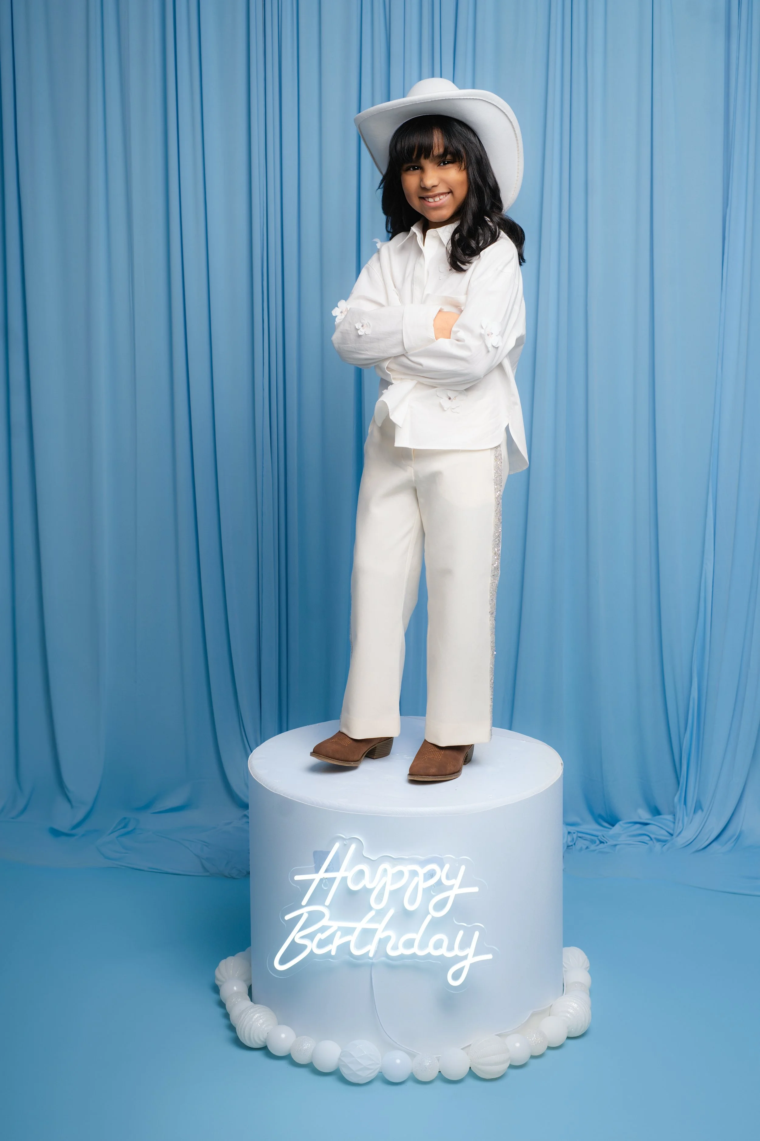 Child dressed in white outfit and cowboy hat standing on a platform with 'Happy Birthday' sign, blue curtain background.