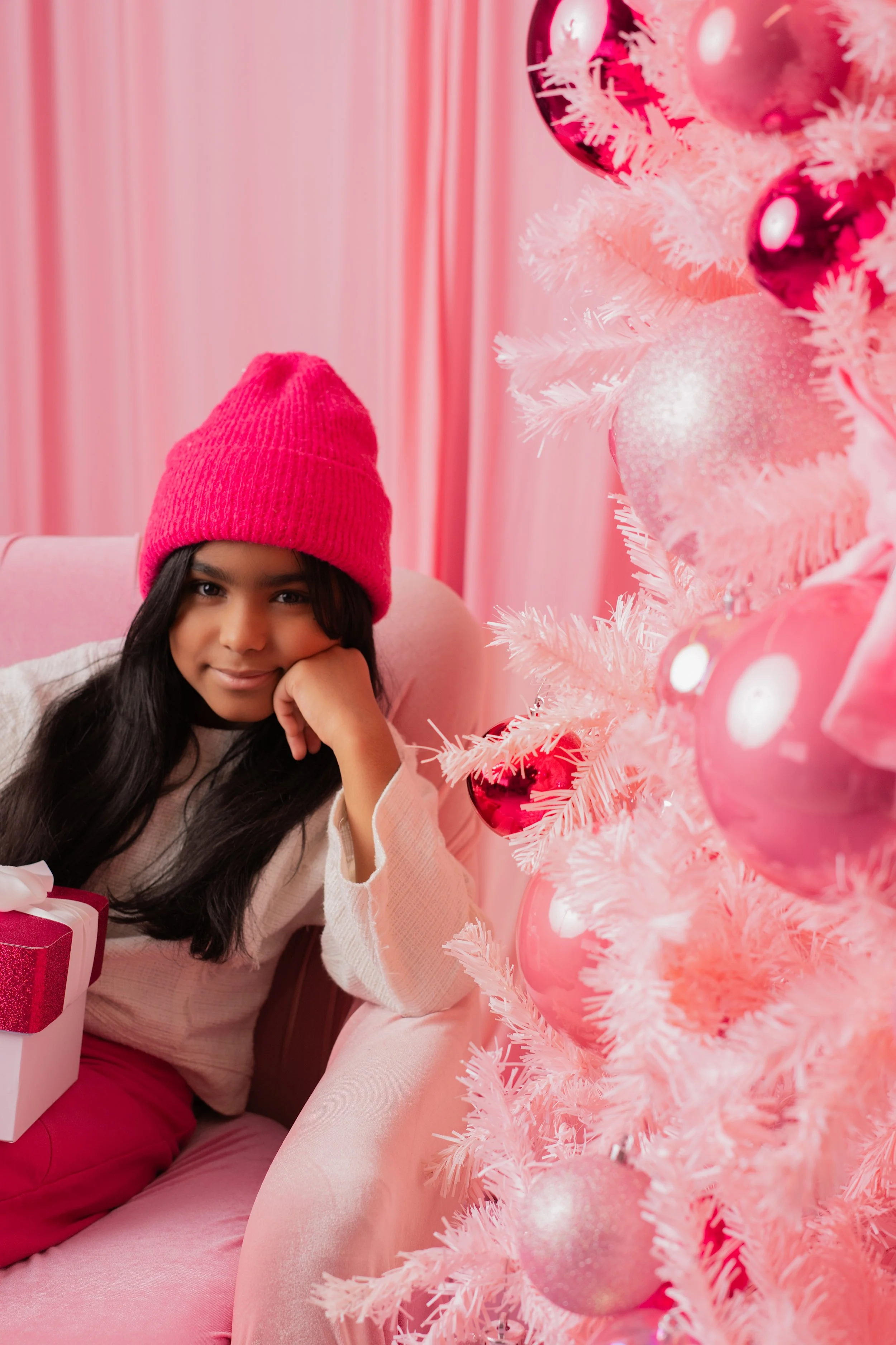 A young girl with long dark hair wearing a pink knit hat, sitting on a pink sofa near a pink Christmas tree decorated with pink and shiny ornaments, holding a wrapped gift box.