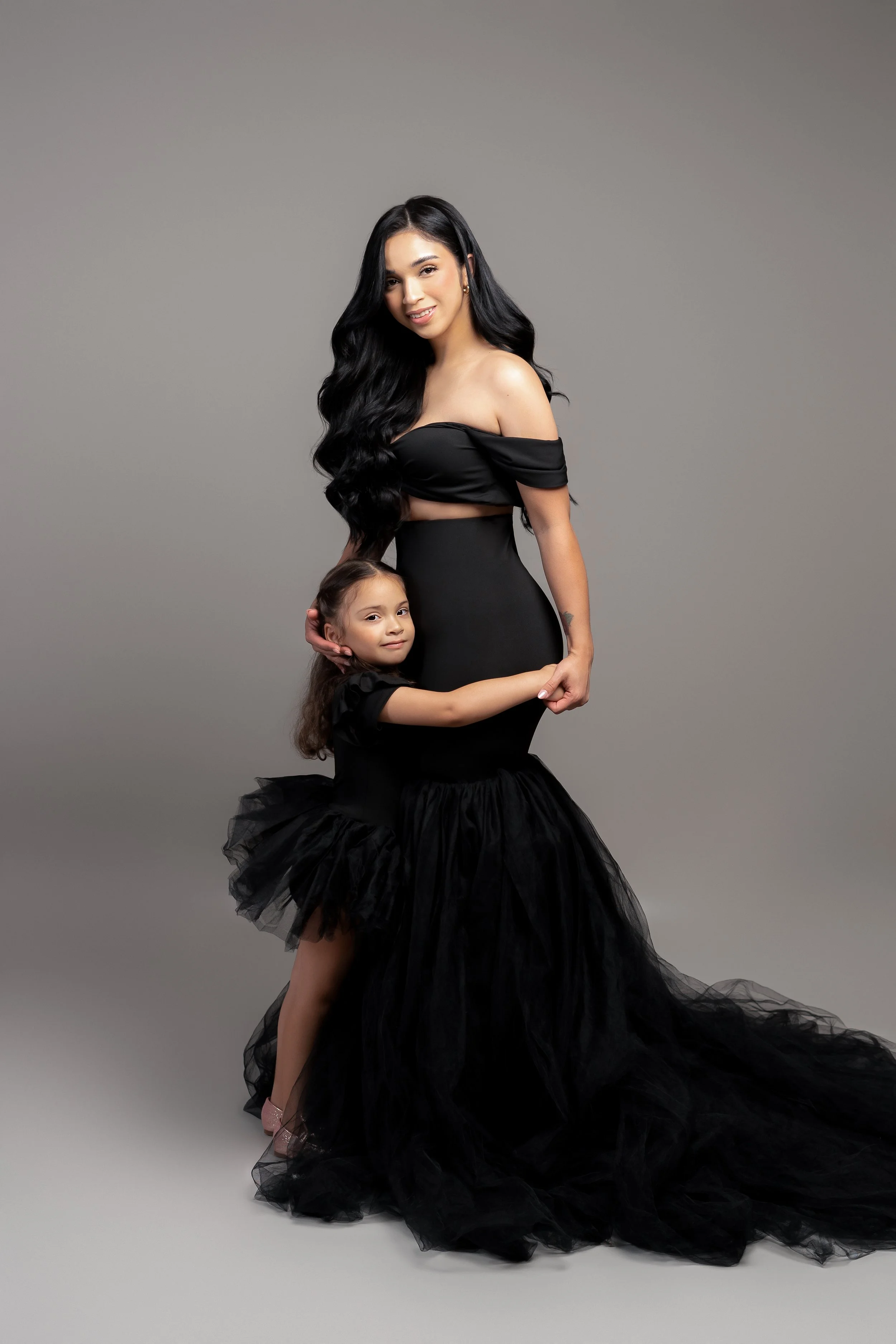 A woman in a black evening gown and a young girl in a matching black dress standing together smiling in a studio setting.