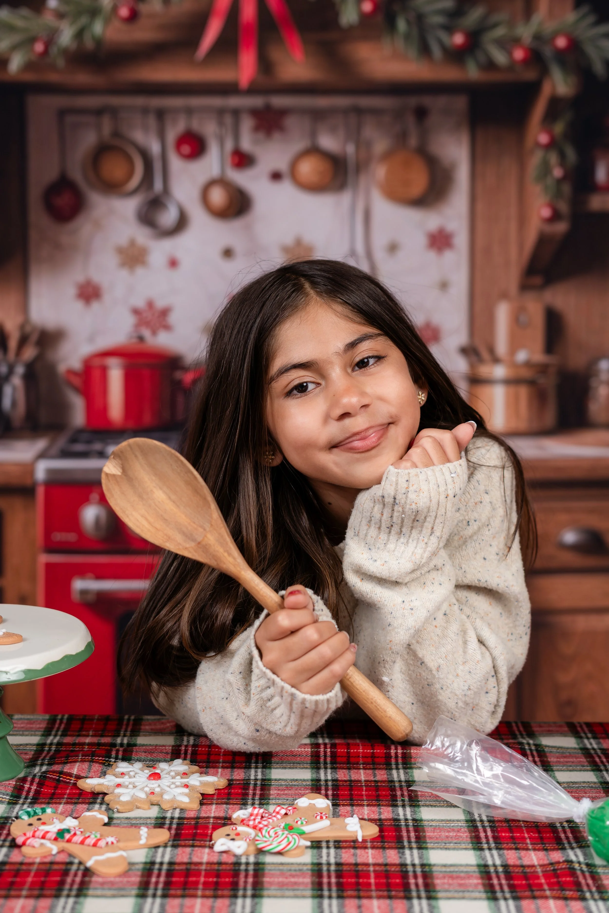 A young girl with long brown hair and a cream-colored sweater smiling at the camera, holding a wooden spoon in a festive kitchen setting decorated for Christmas, with decorated cookies on the table in front of her.