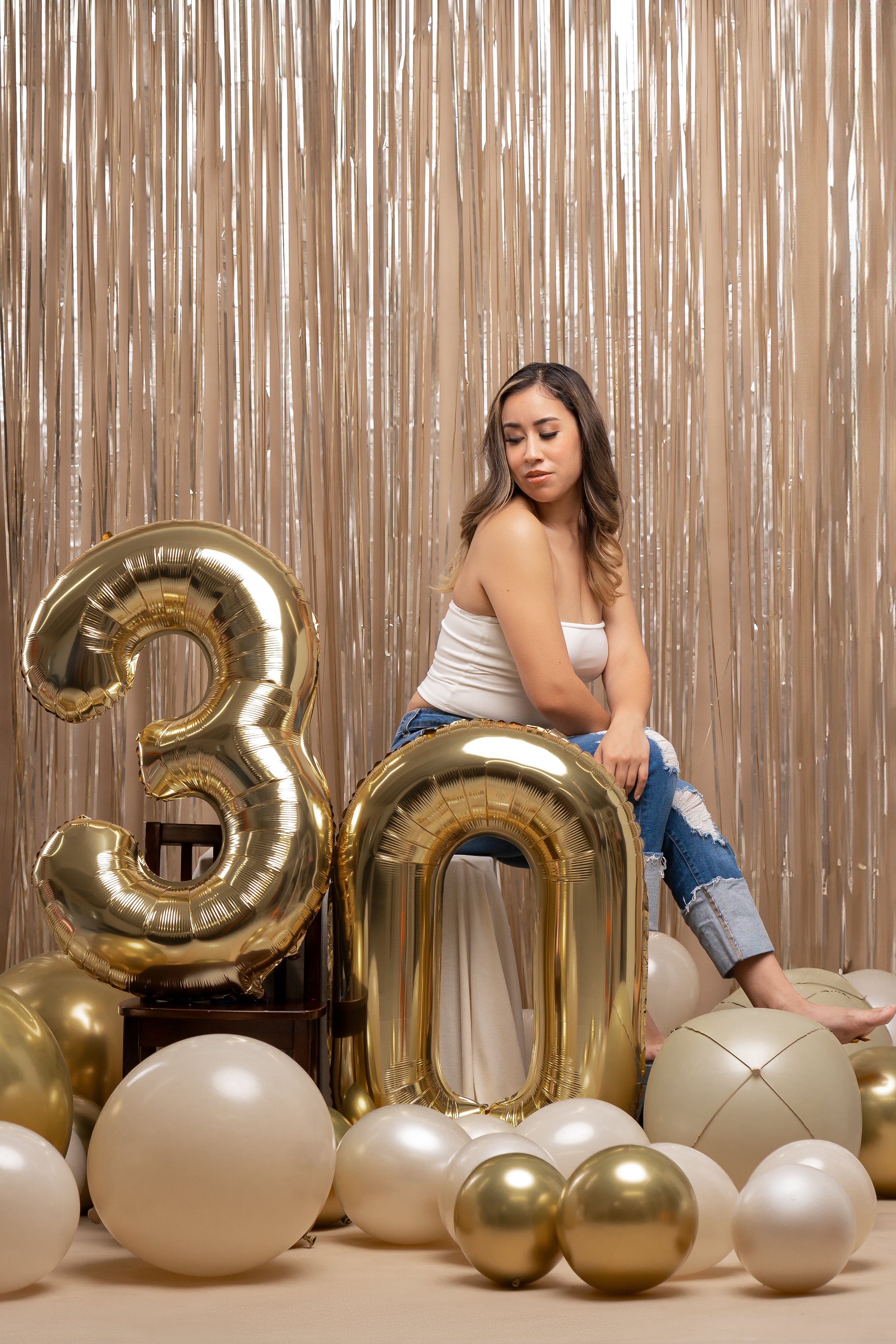 A woman sitting in front of gold foil curtains, surrounded by gold and white balloons, with large gold balloons forming the number 30.