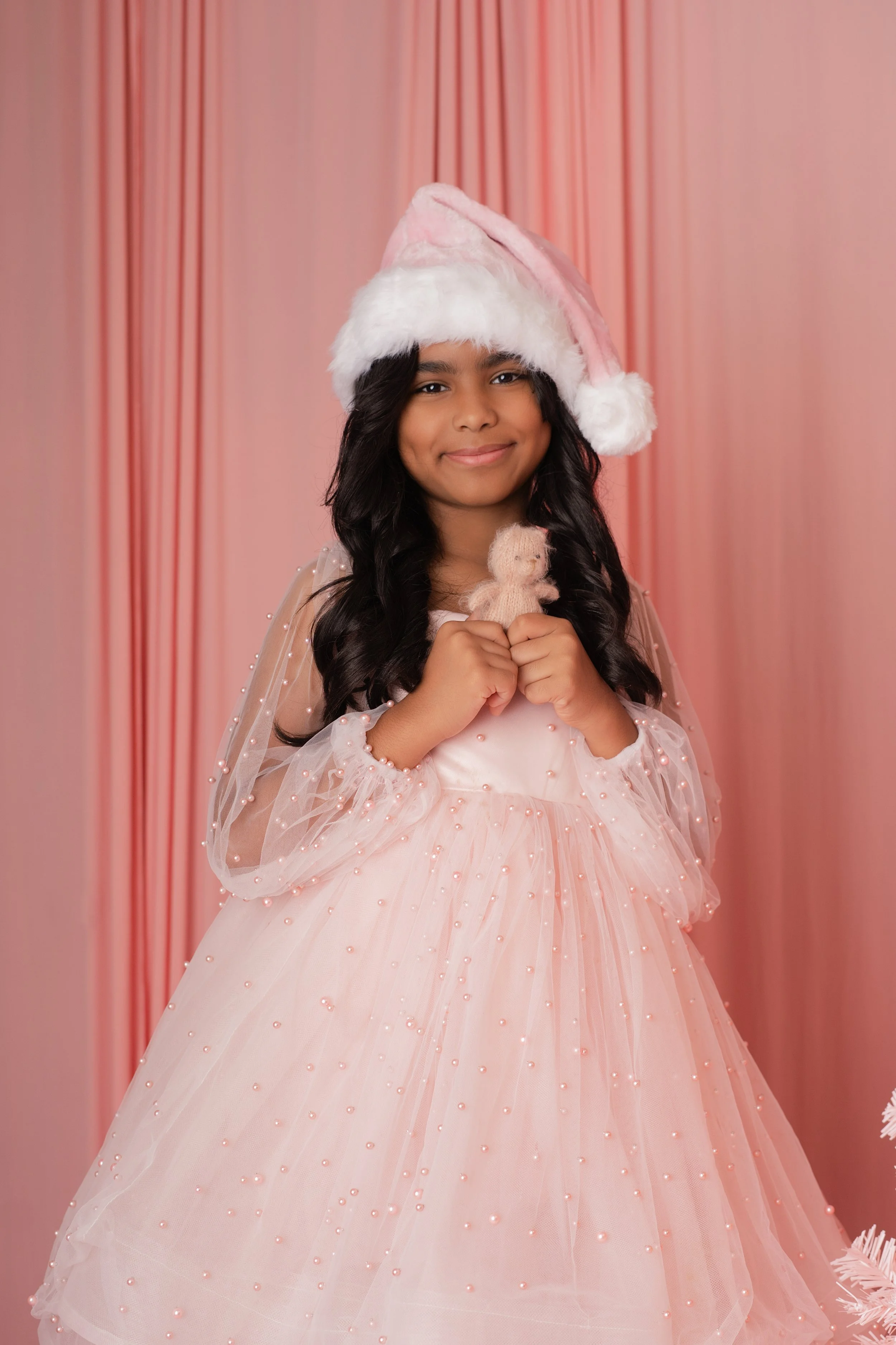 A young girl dressed in a pink Christmas dress with pearl embellishments, wearing a Santa hat, holding a small stuffed bear, standing against a pink curtain background.