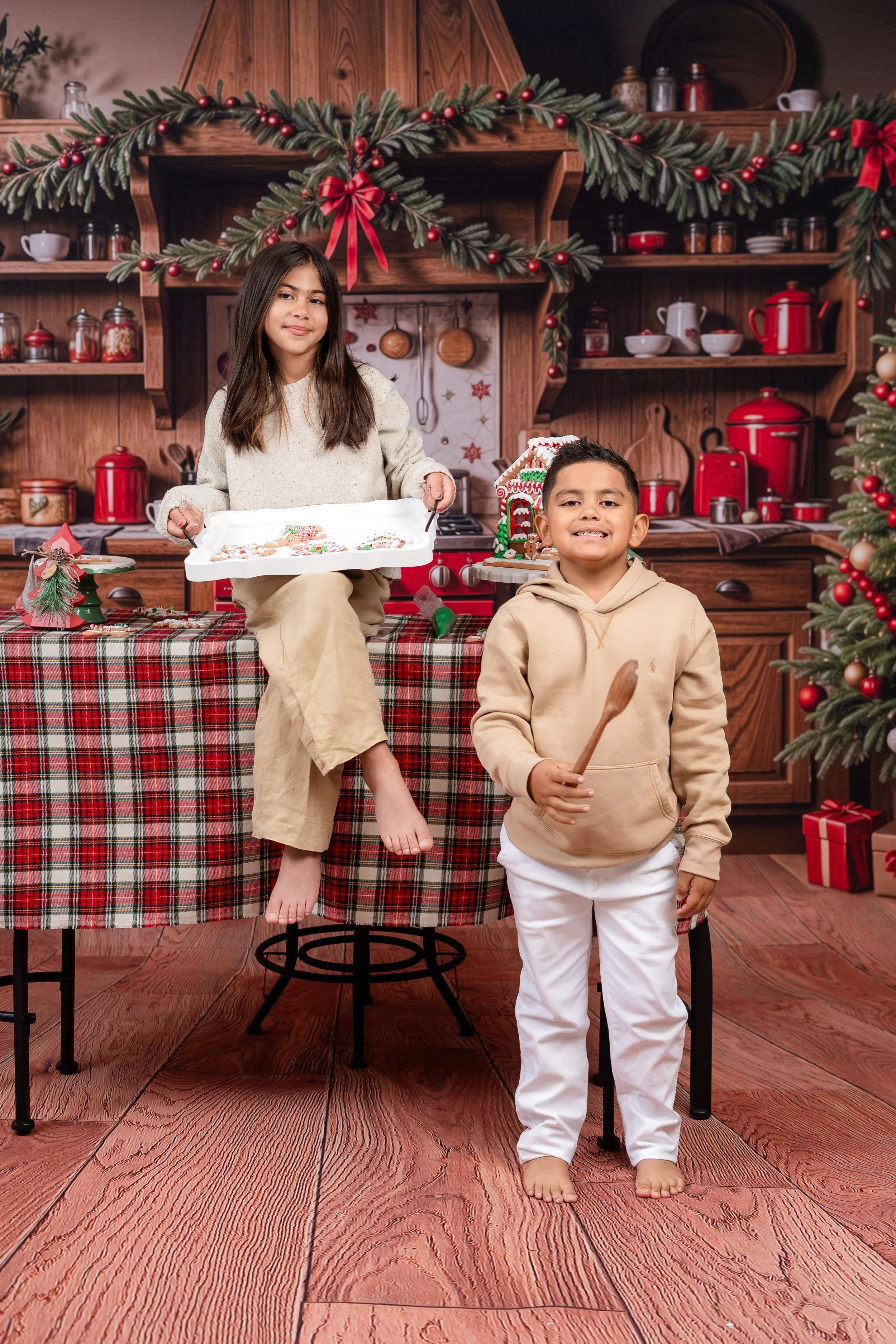 Two children in a decorated kitchen for Christmas, with a girl sitting on a table holding a tray of decorated cookies and a boy standing next to her holding a spoon, both smiling.