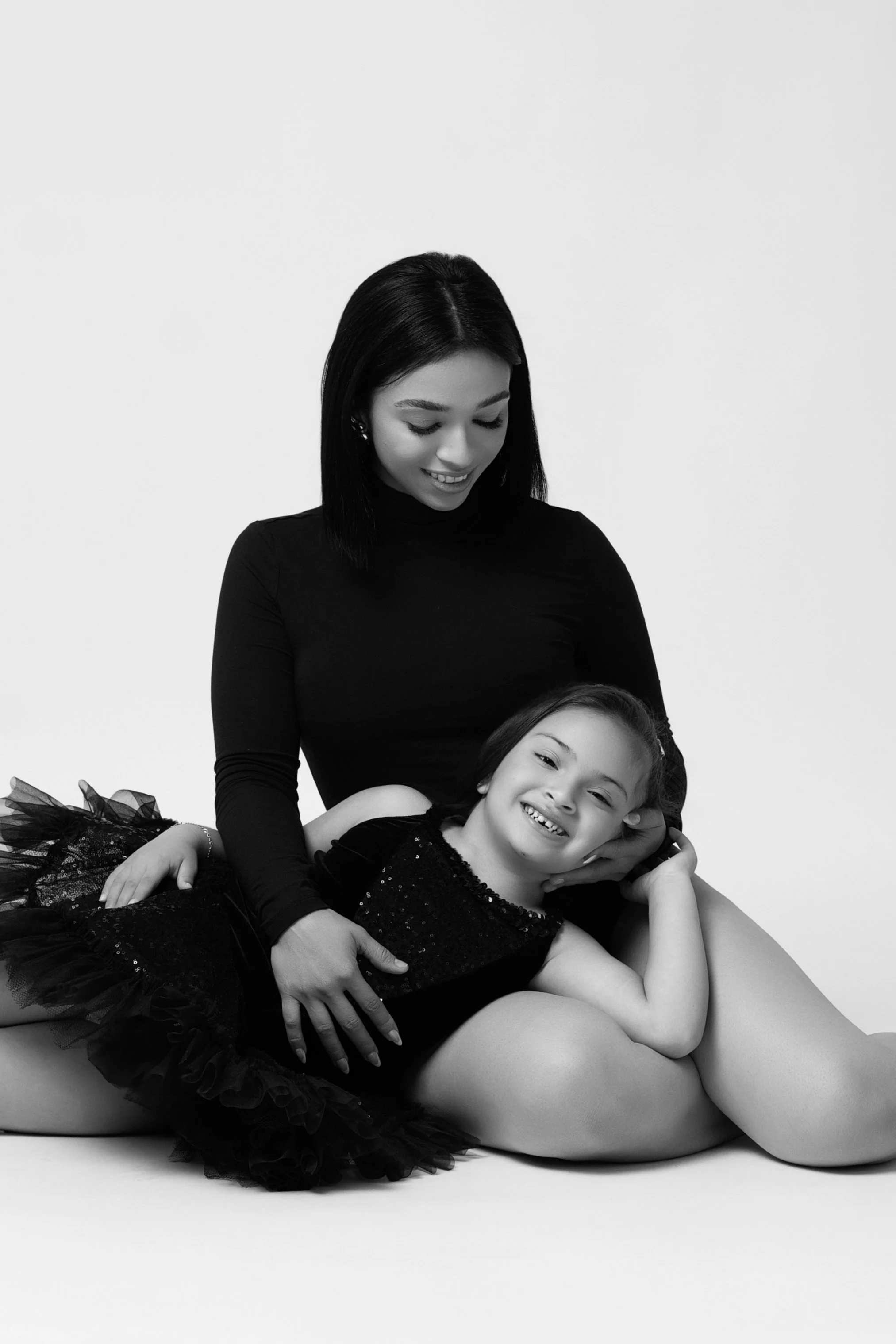 Black and white photo of a smiling mother and daughter sitting together, both wearing black outfits, the daughter leaning on the mother's lap.