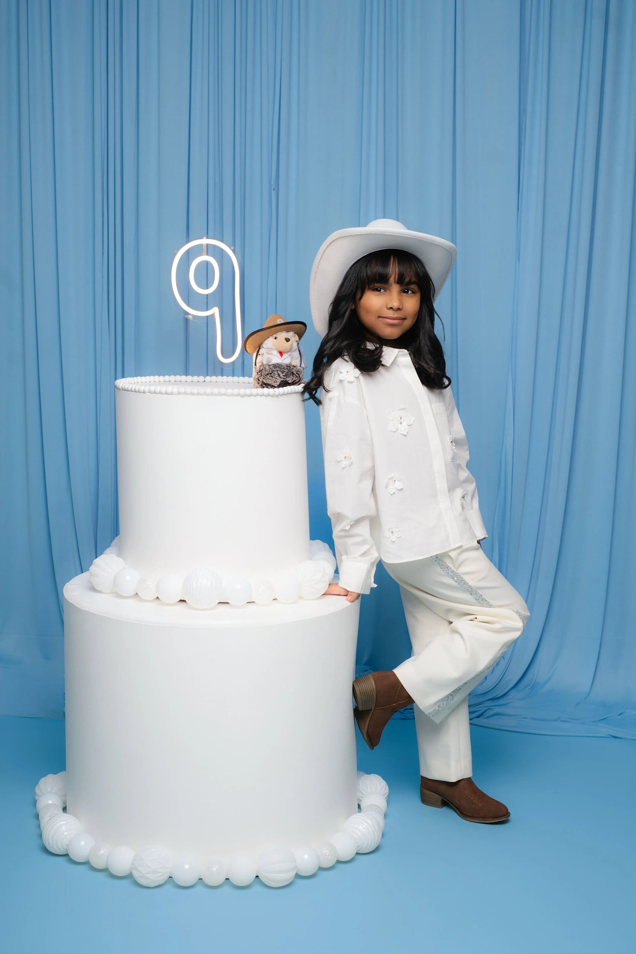 Child wearing a white cowboy hat and outfit next to a large cake with a neon number "9" and a toy in a cowboy hat on top, against a blue backdrop.