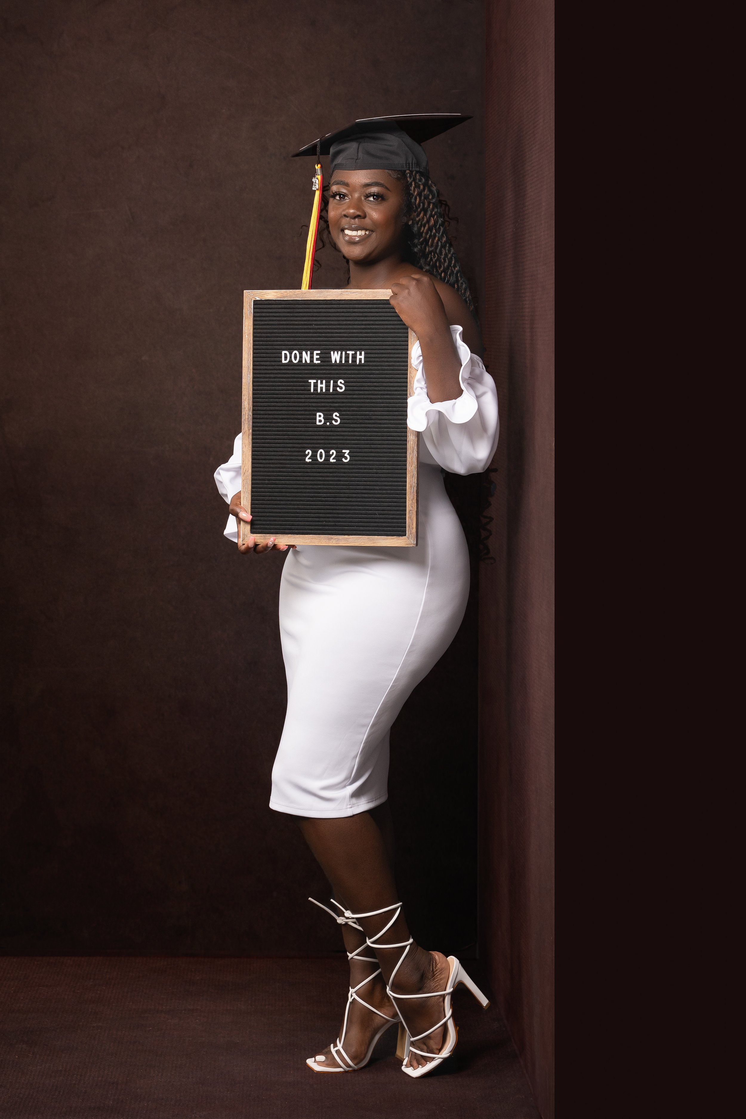 Woman wearing a graduation cap and white dress, holding a letter board with "Done with this B.S 2023" written on it, and wearing strappy heels.