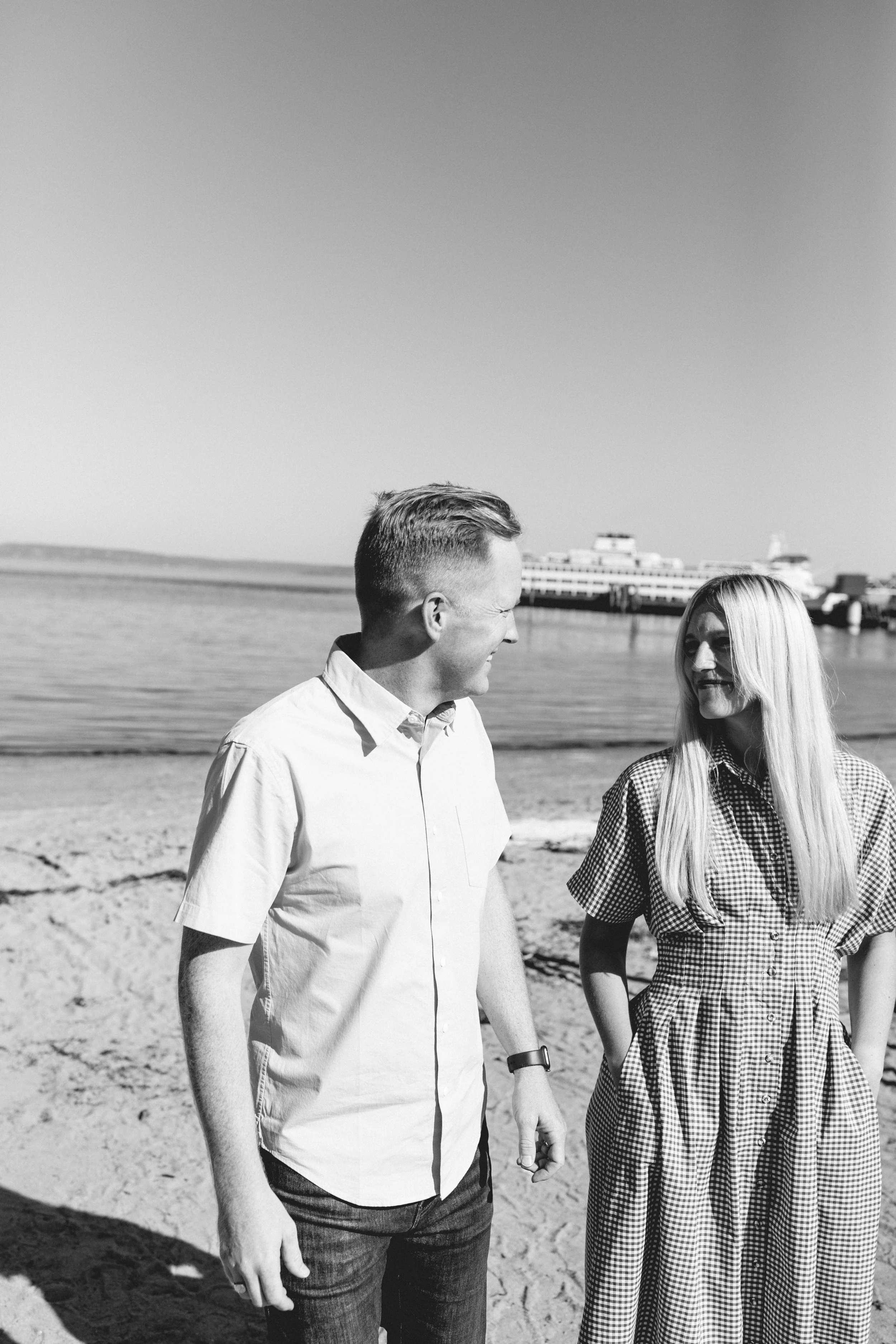 A black and white photo of a man and woman standing on a beach by the water, smiling and looking at each other.