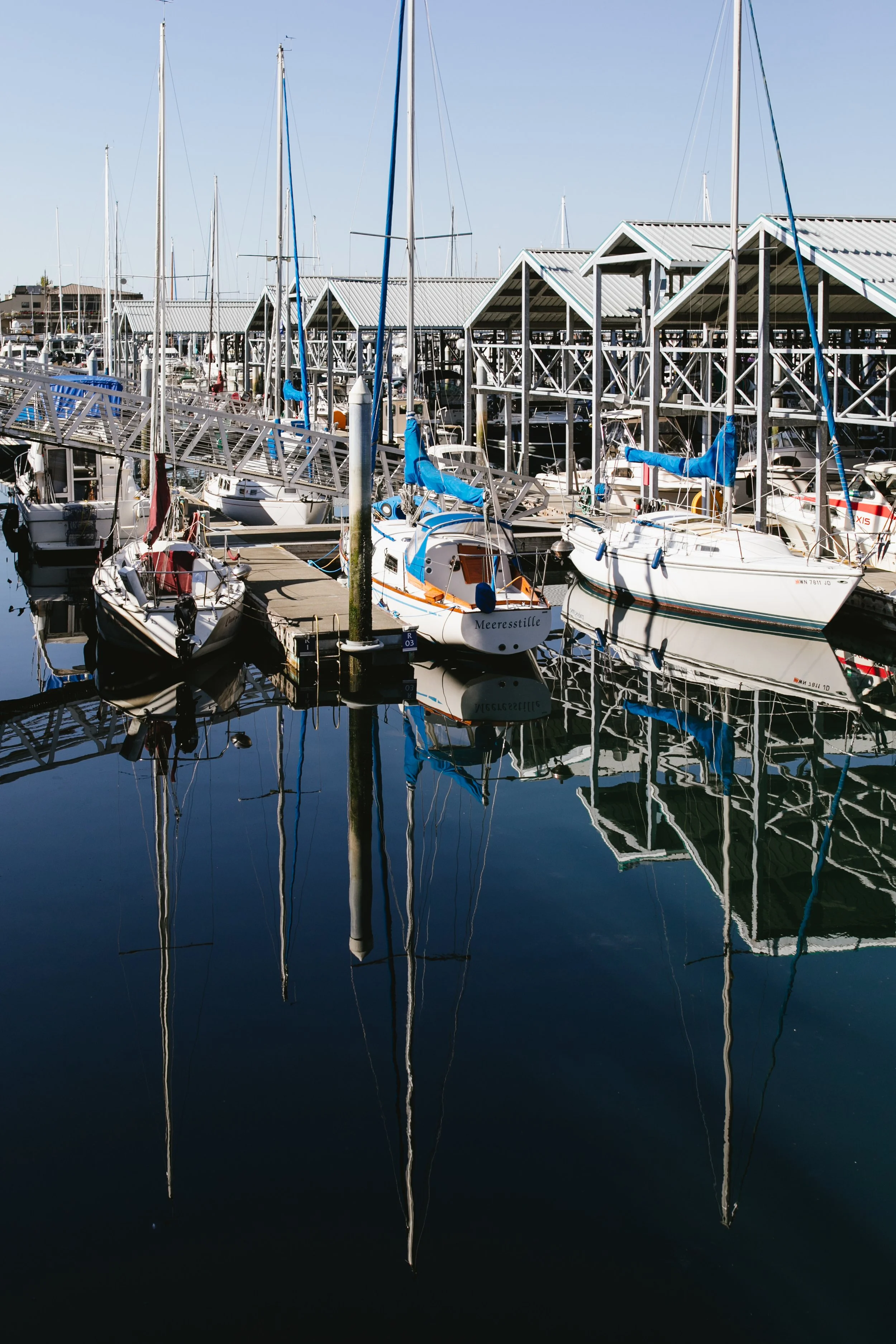 Marina with sailboats docked, wooden docks, and covered racks for boats, with their reflections visible in the calm water.