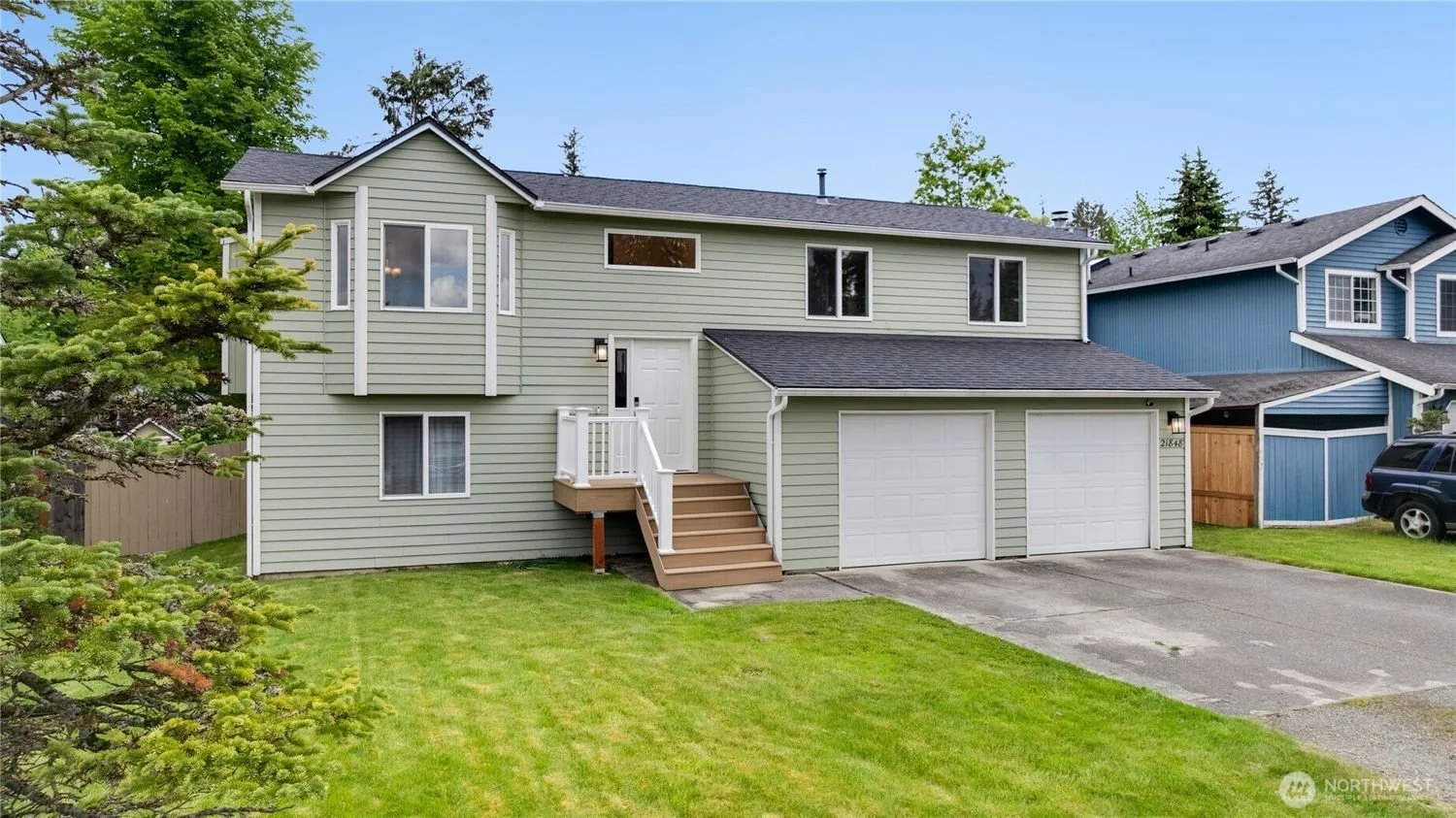 A two-story house with light green siding, a garage with two doors, and a small front staircase with white railing. The house is surrounded by a green lawn and trees, with neighboring houses visible in the background.