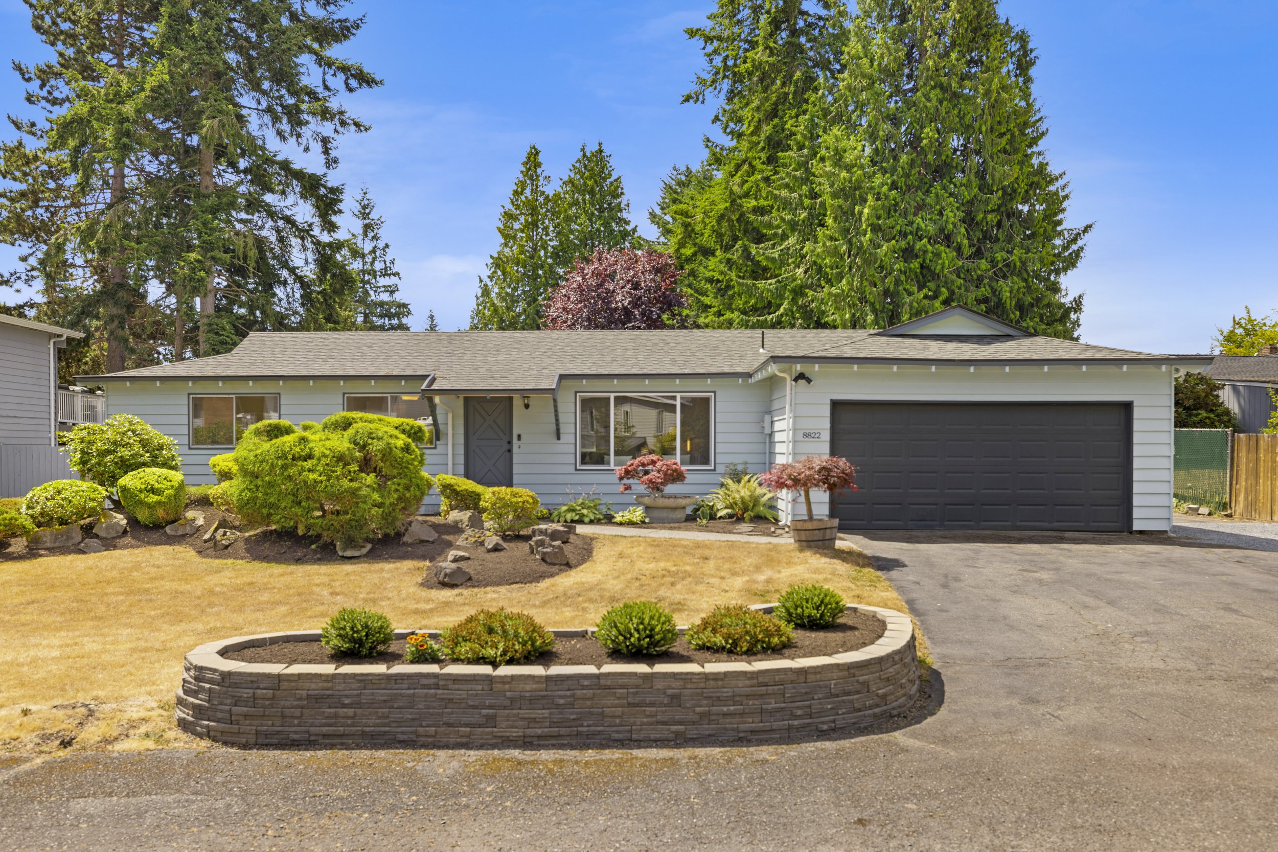 Single-story suburban house with a gray exterior, attached garage, and front yard with landscaping including bushes, small trees, and a curved stone planter wall, under a blue sky with large trees in the background.