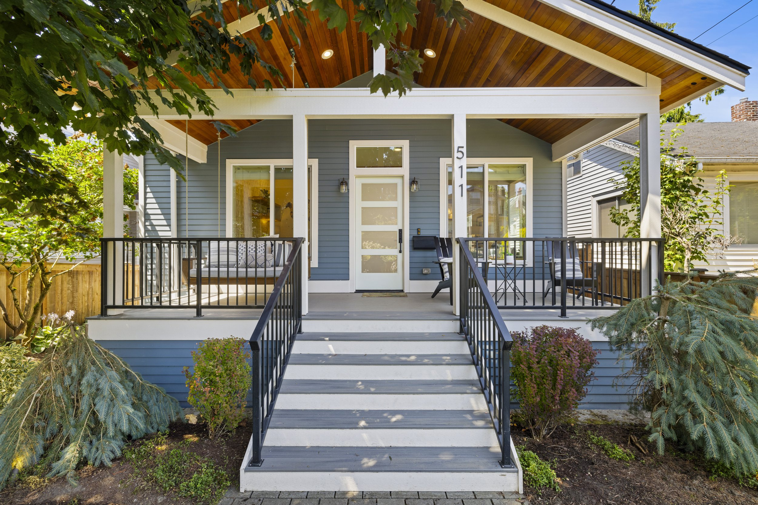 Front porch of a modern house with blue exterior, white trim, and black railing, accessible by a short staircase, with porch furniture and greenery.