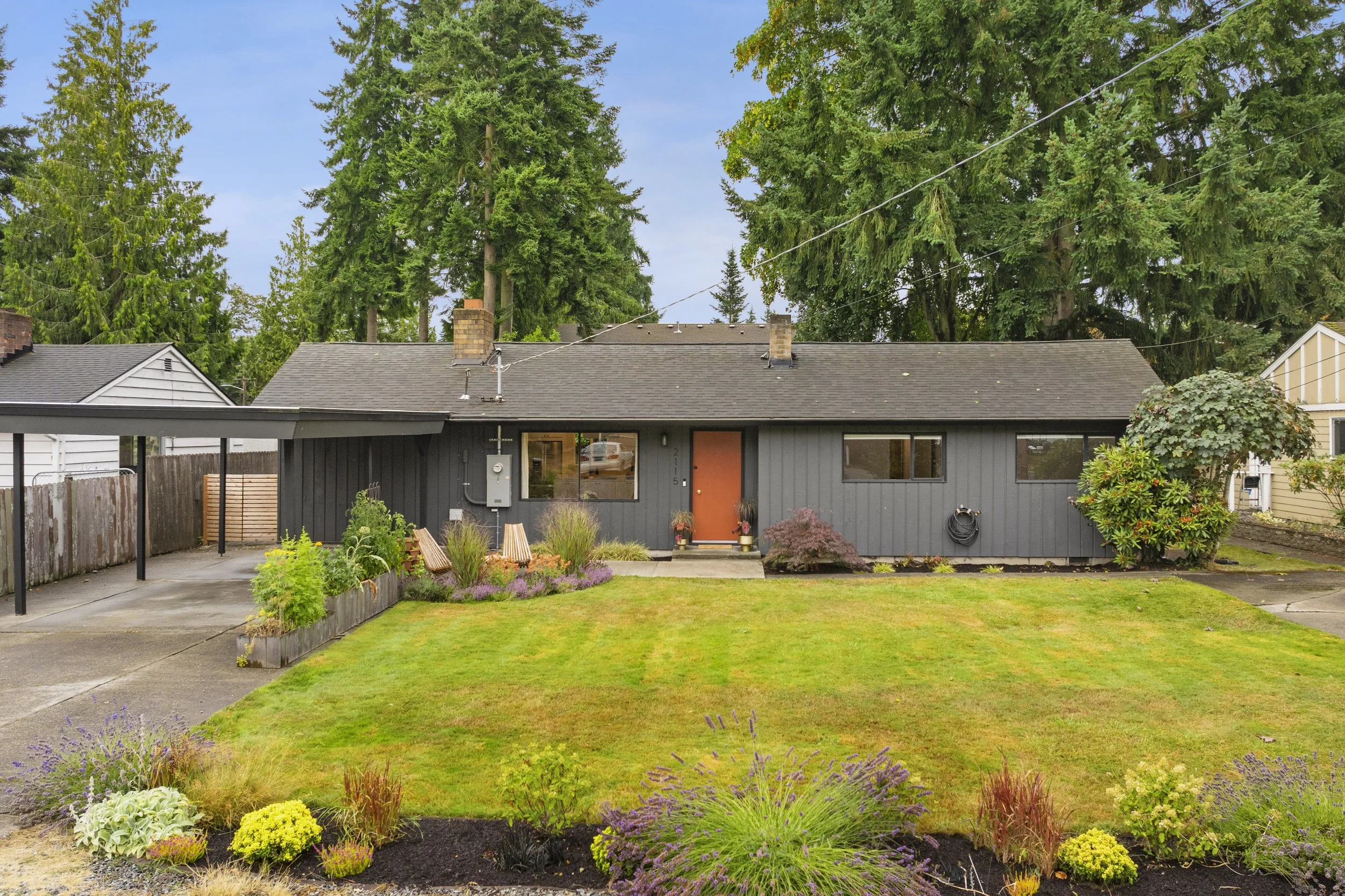 Front yard of a single-story house with a grassy lawn, flower beds, and large trees in the background.