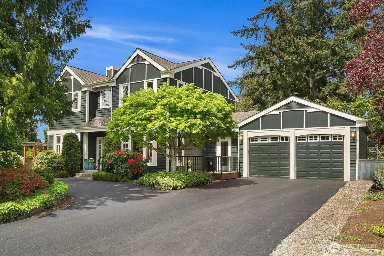 A modern two-story house with black and white exterior, surrounded by green trees and plants, and a paved driveway leading to a double garage.