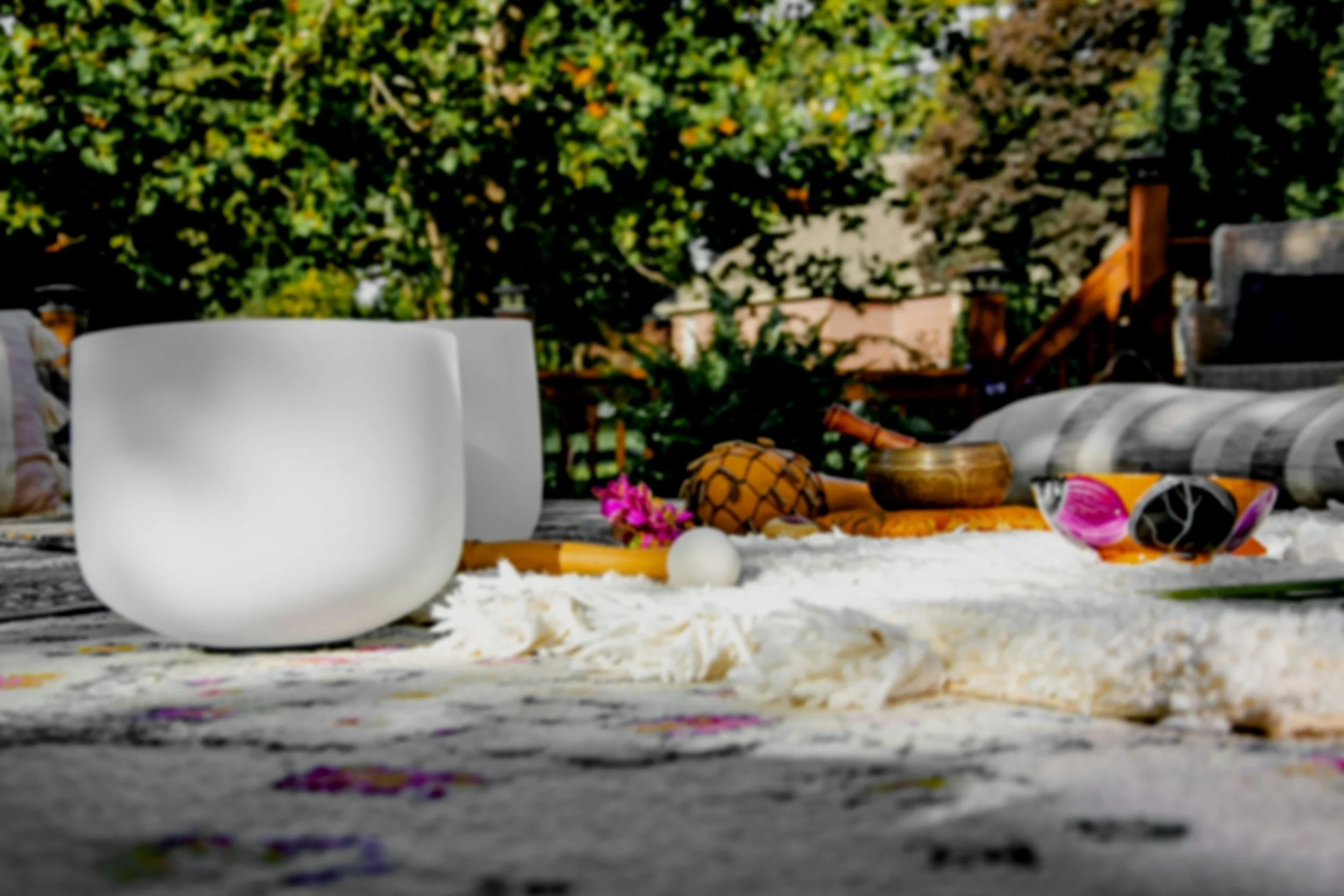 Outdoor scene with singing bowls, a pineapple, flowers, and a cloth on a mat, set against a background of green trees and a wooden deck.