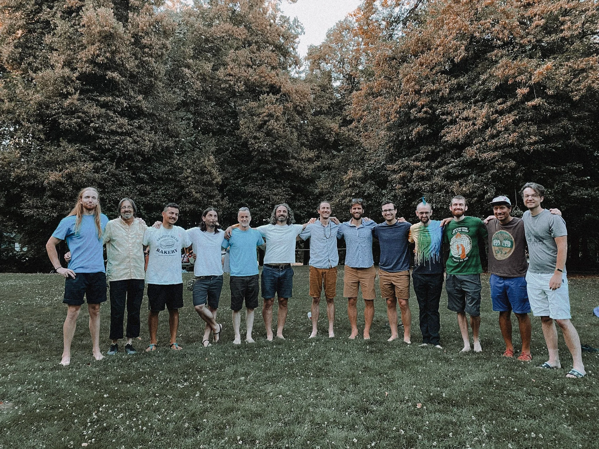 A group of fifteen men standing outdoors on grass with trees in the background, arms around each other's shoulders, smiling.