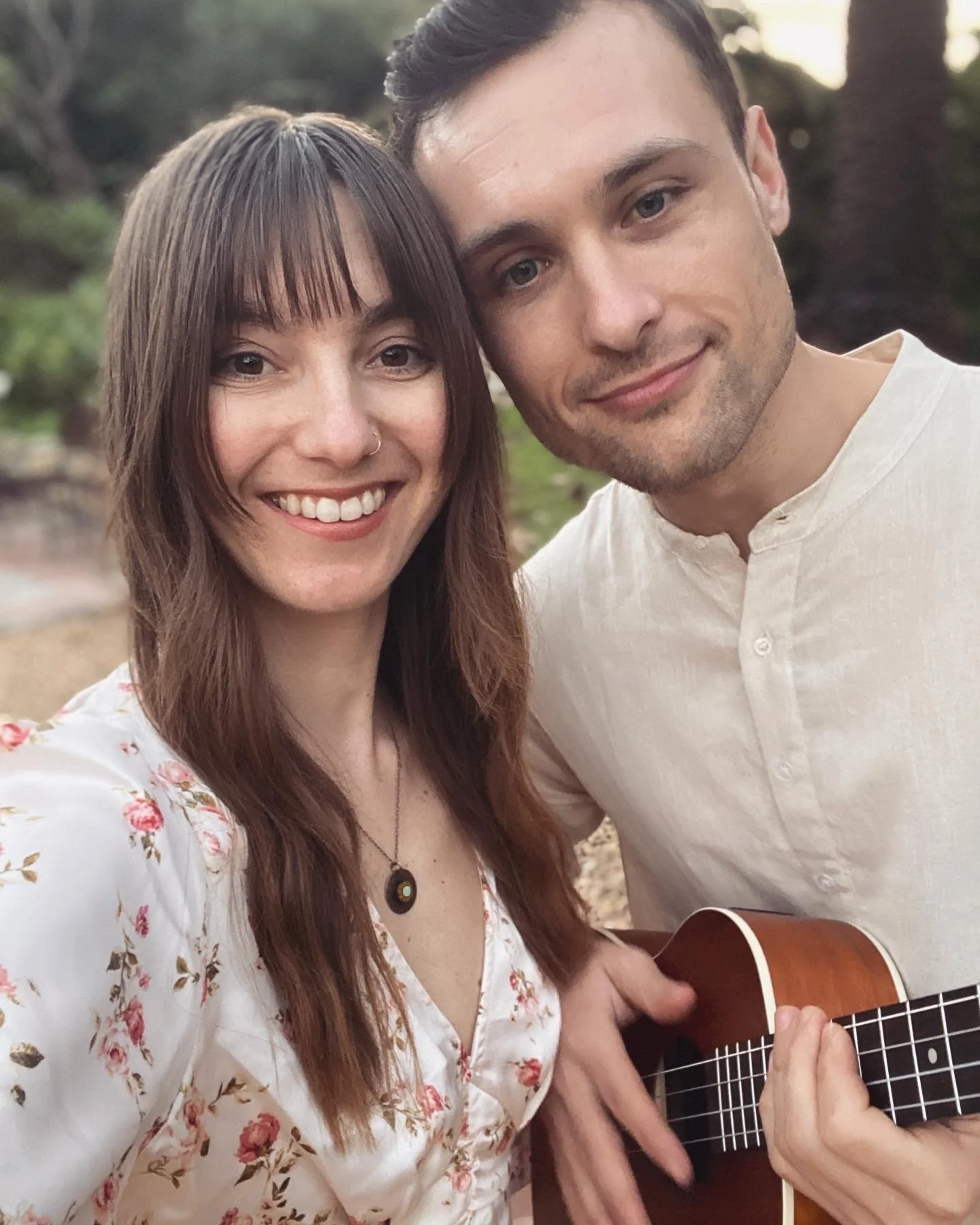 A smiling woman with long brown hair holding a guitar and a man with short dark hair, both outdoors in a natural setting.