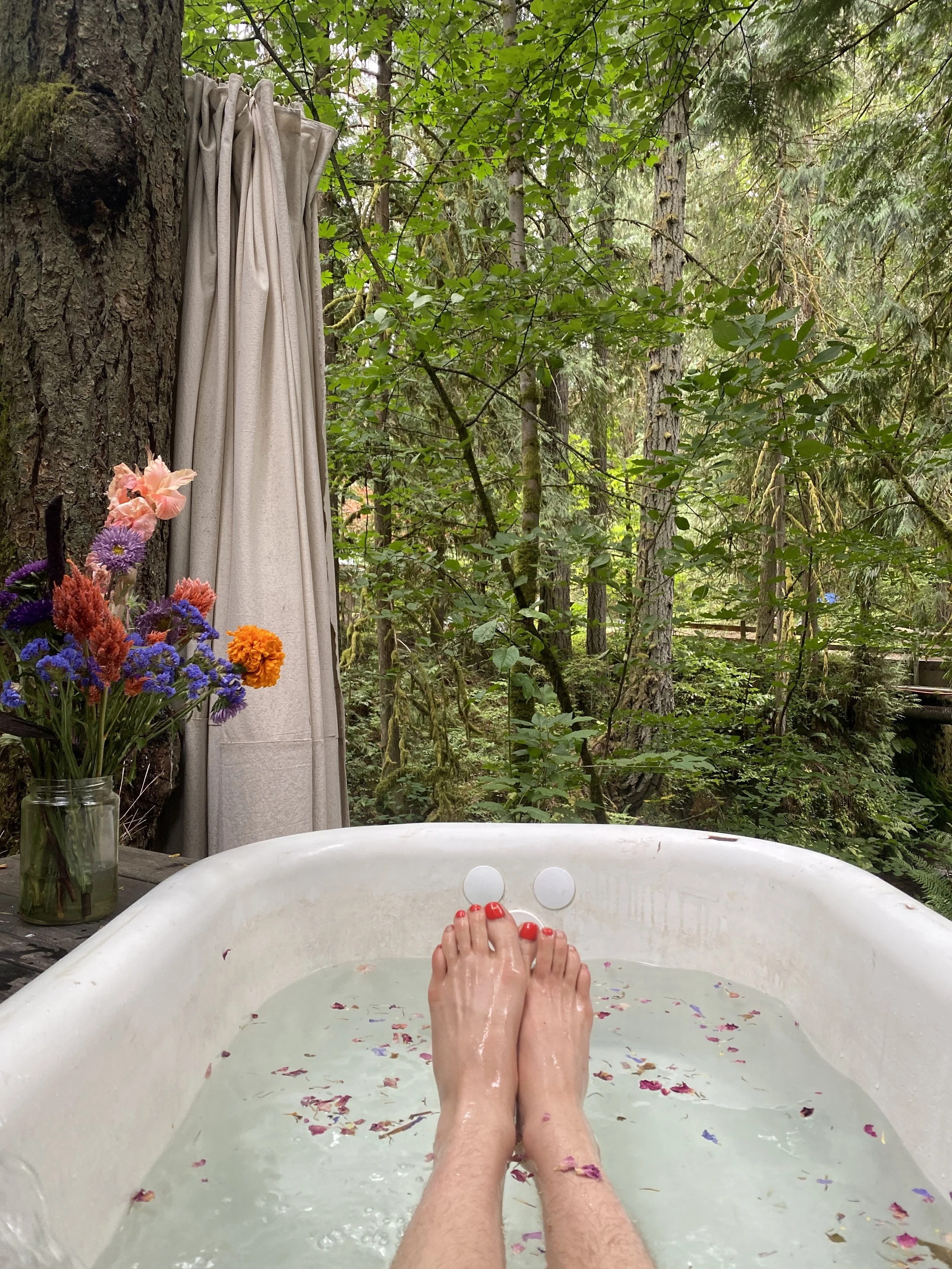 A person with red nail polish is soaking in an outdoor bathtub filled with water and flower petals, set in a lush green forest with trees and foliage. A jar of colorful flowers and a curtain are visible nearby.